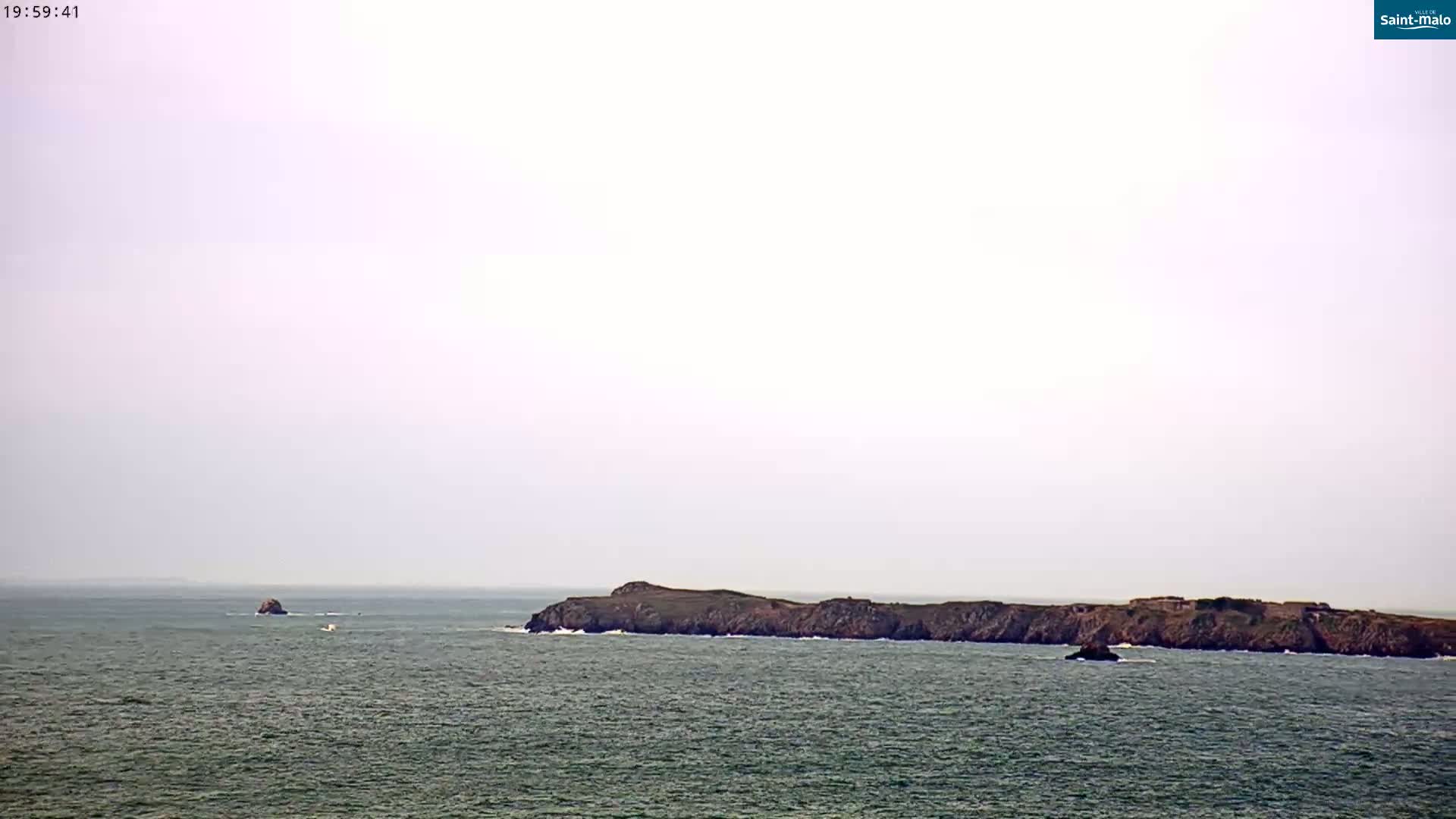 A long, low-lying island is visible on a calm, overcast day, with a smaller rock formation further out to sea.