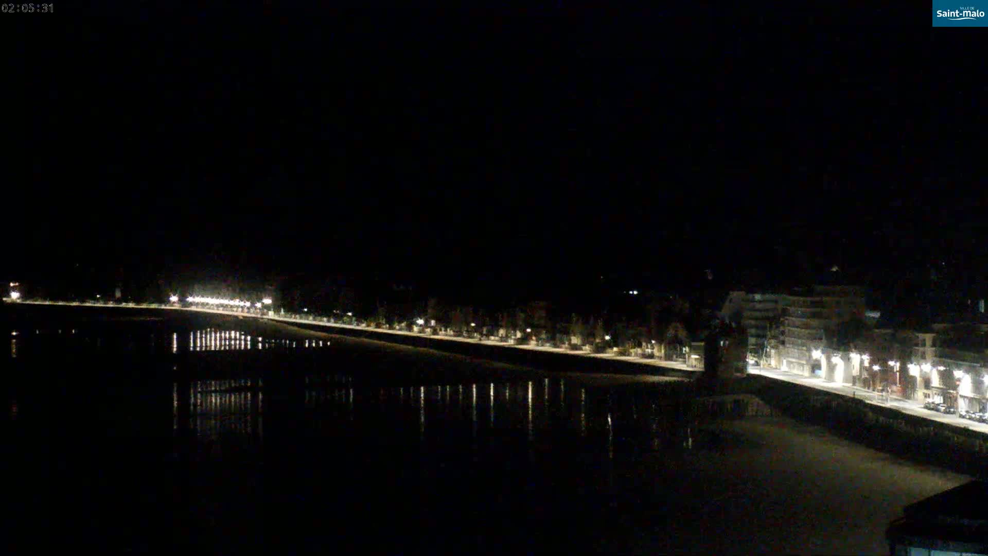 A nighttime, long shot of a waterfront town with lights reflecting on calm water under a clear, dark sky.
