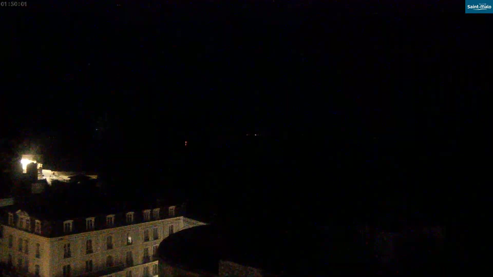 A dark nighttime scene shows a portion of a building illuminated by interior lights, with faint distant lights visible in the darkness.