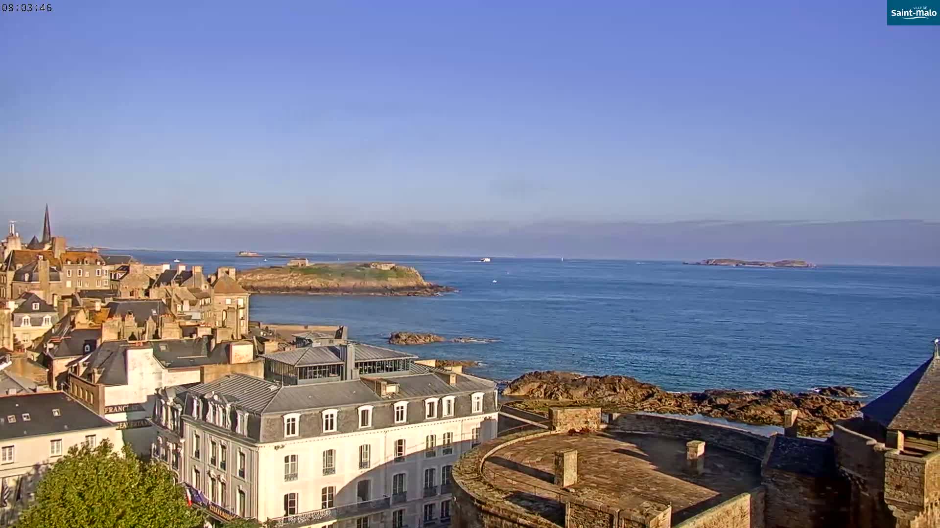 A coastal town with stone buildings overlooks a calm blue ocean and several small islands under a clear blue sky.