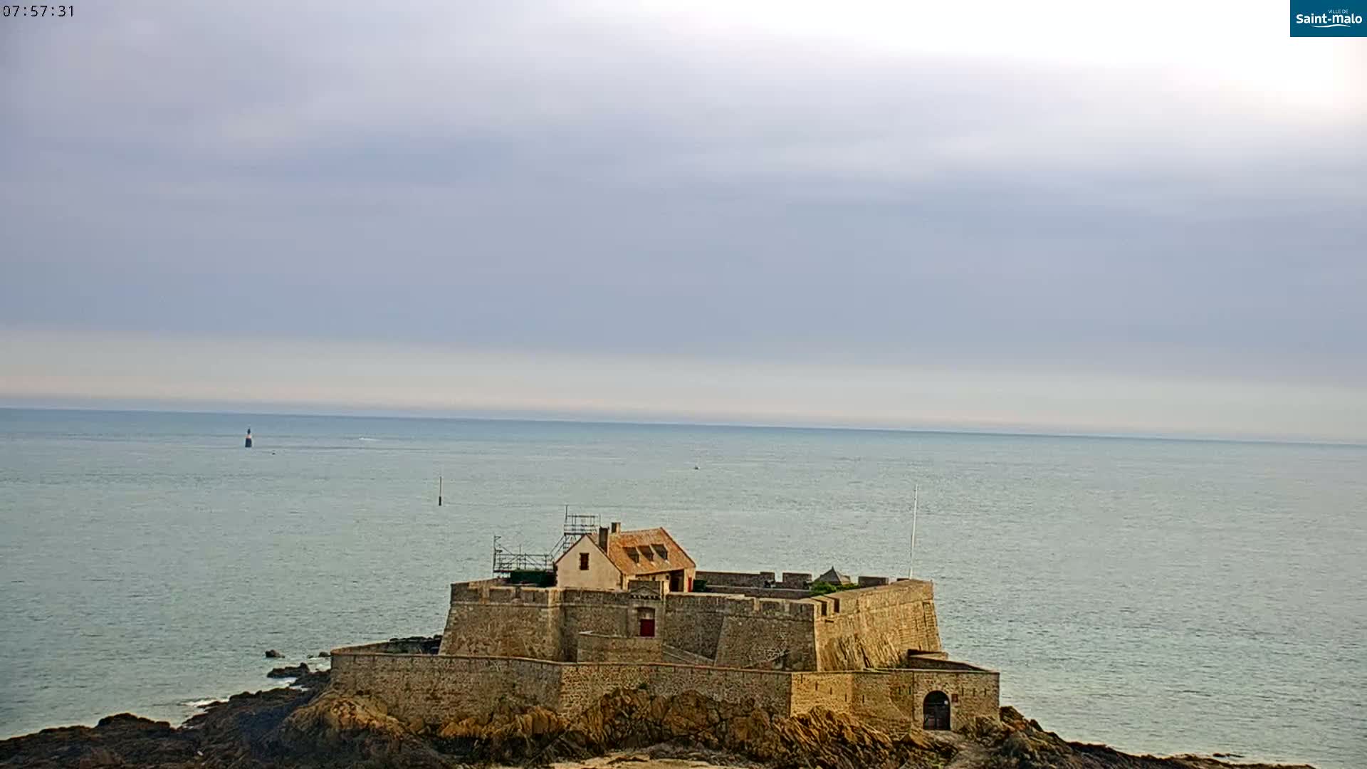 A stone fort with a small house sits on a rocky outcrop jutting into a calm sea under a cloudy sky.