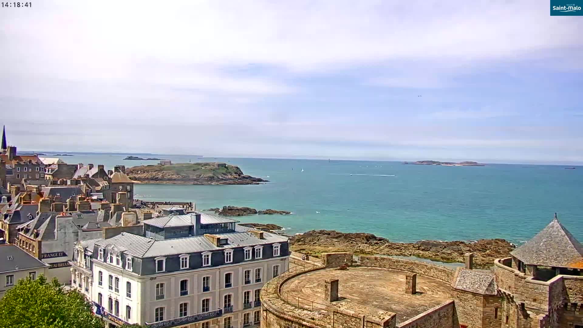 A coastal city with stone buildings overlooks a calm, turquoise ocean dotted with small islands under a partly cloudy sky.