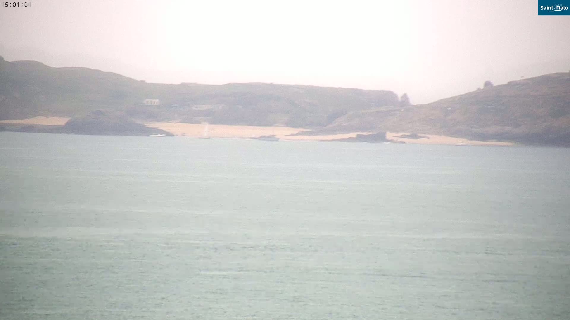 A hazy view shows a calm body of water with a distant shoreline featuring low-lying hills and a small sandy beach.