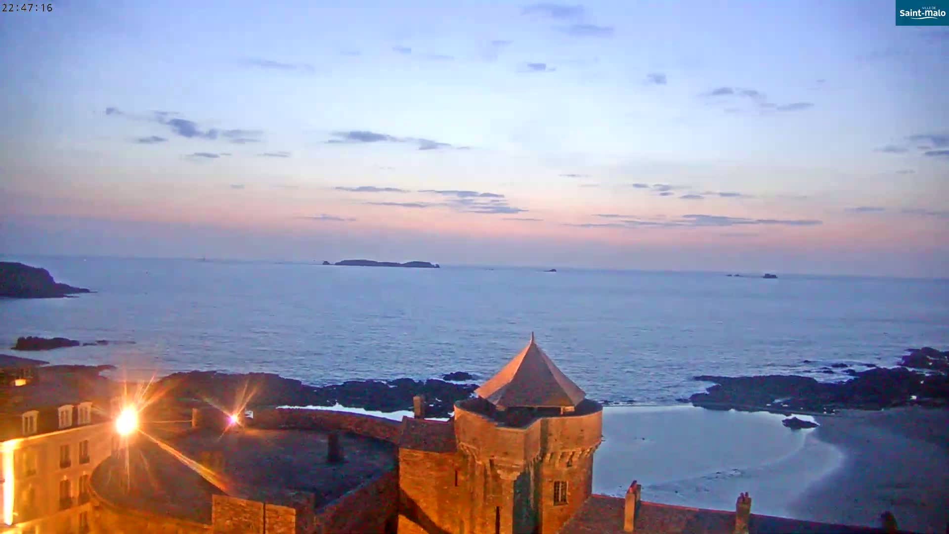 A coastal scene at twilight shows a stone tower overlooking a calm ocean with a small island in the distance under a pale pink and blue sky.