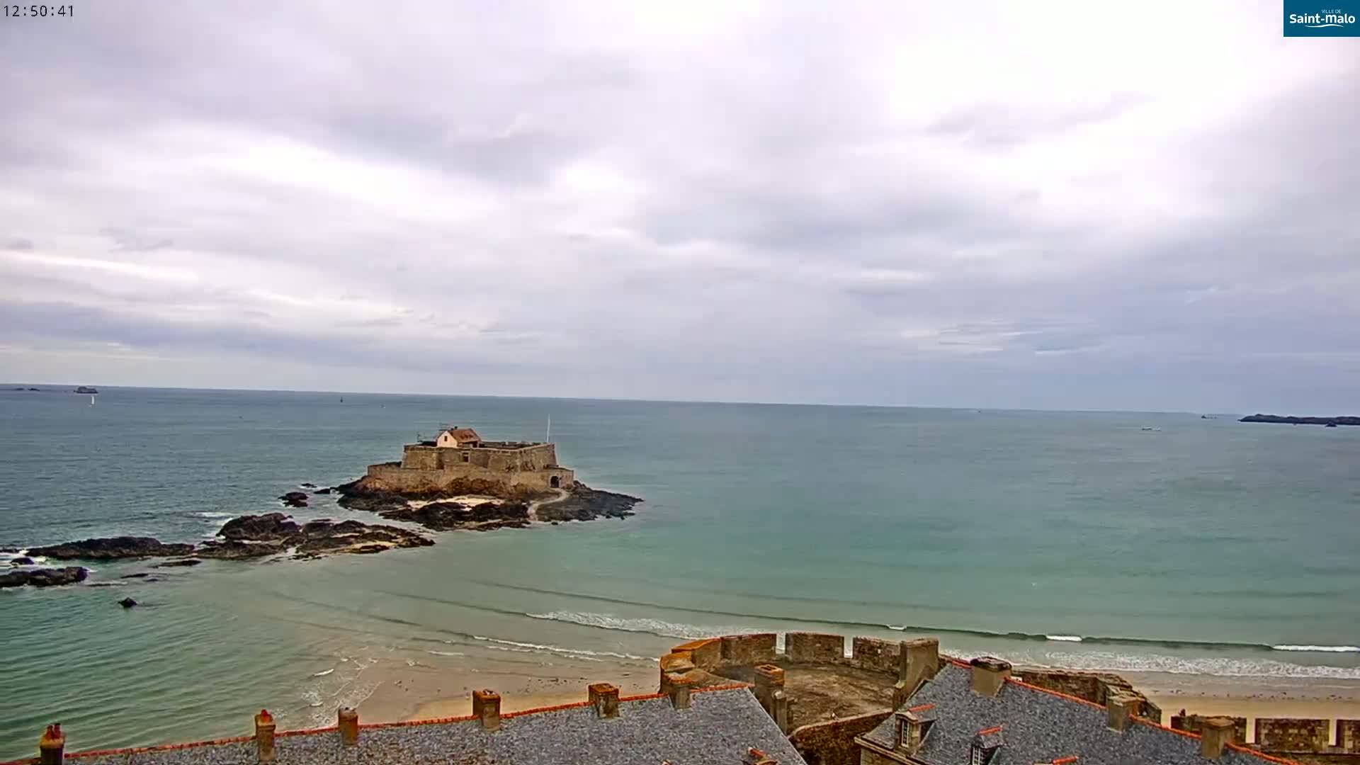 A coastal scene under a cloudy sky shows a stone castle on a small island in a calm sea with a sandy beach and town rooftops in the foreground.