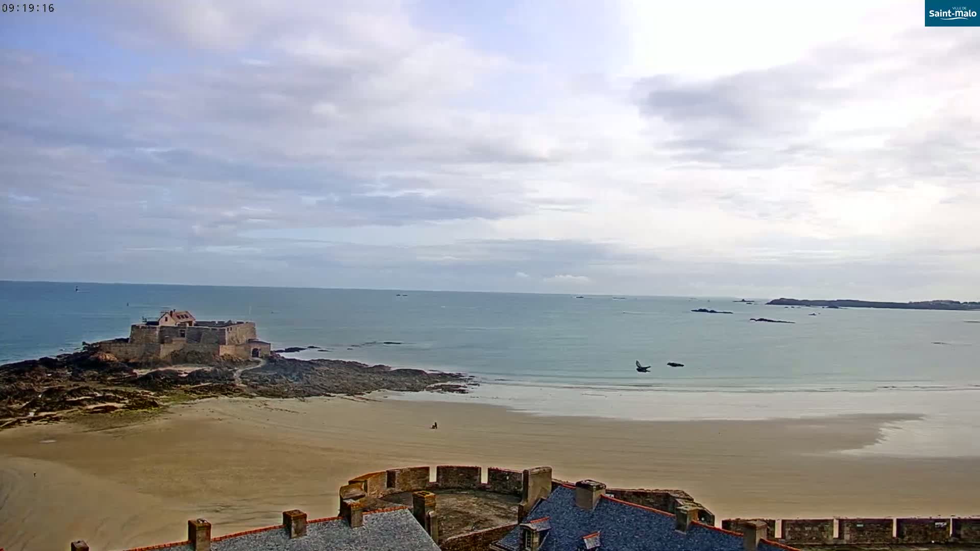 A stone castle sits on a rocky outcrop connected to a sandy beach by a narrow strip of land, under a mostly cloudy sky.