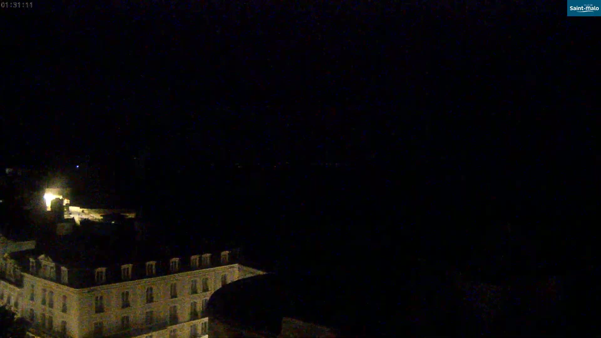 A dark nighttime view shows a portion of a multi-story building illuminated by faint interior lights, overlooking a mostly obscured landscape.