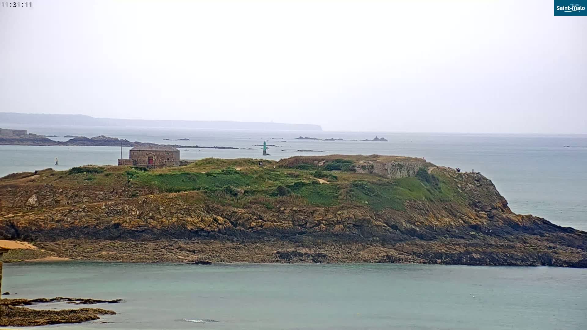 A rocky island with a stone building and a lighthouse, surrounded by calm, grey-blue water under an overcast sky.