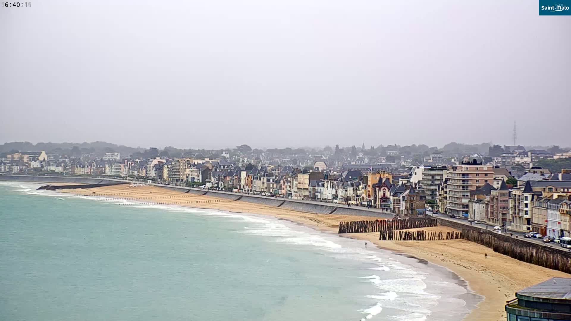 A coastal town with a sandy beach and calm, light-green ocean under an overcast sky.