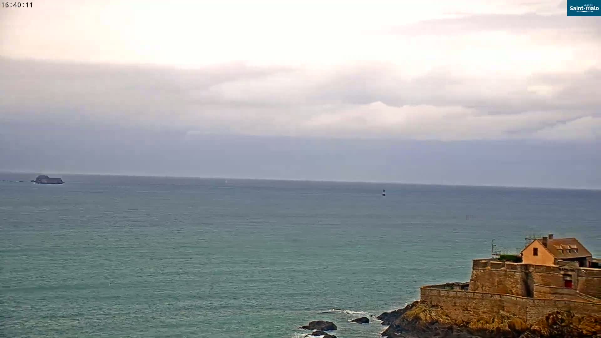 A stone coastal fortification with a small building on top overlooks a calm, overcast ocean with a small island in the distance.