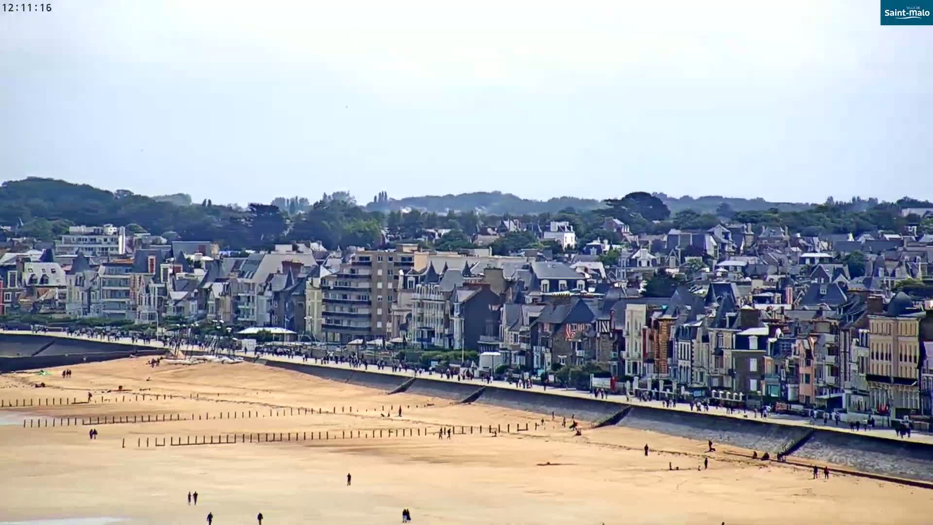 A coastal town with a sandy beach and many people walking along the shore on a clear, sunny day.