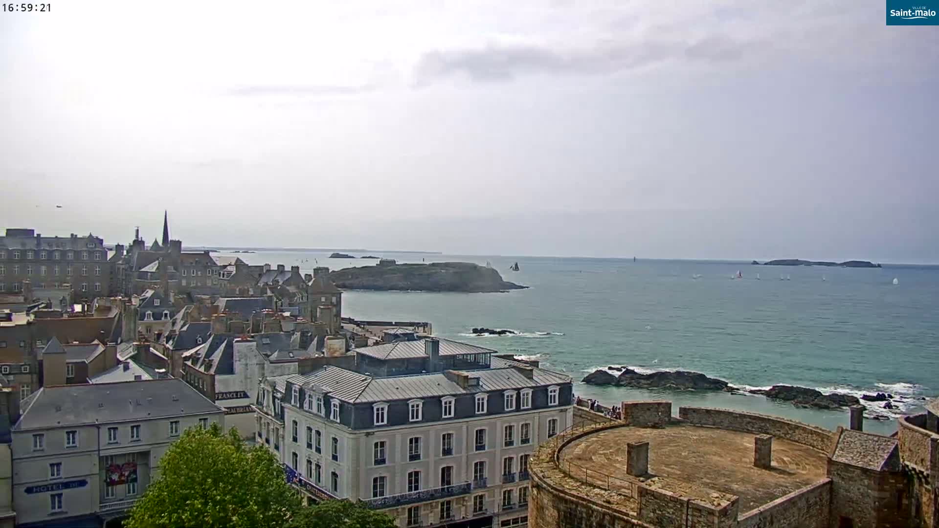 A coastal city with stone buildings overlooks a calm sea dotted with sailboats under a mostly cloudy sky.