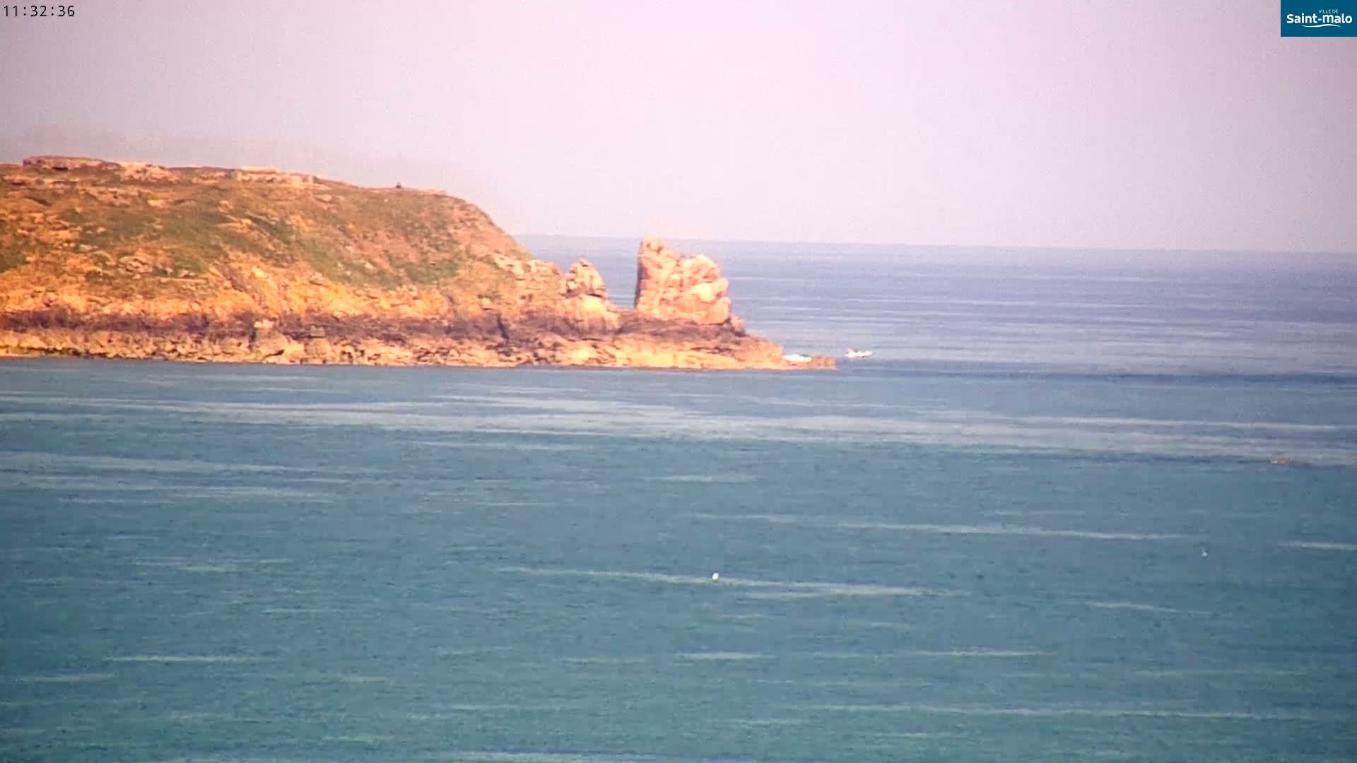 A rocky, grassy headland juts into a calm, blue ocean under a hazy sky.