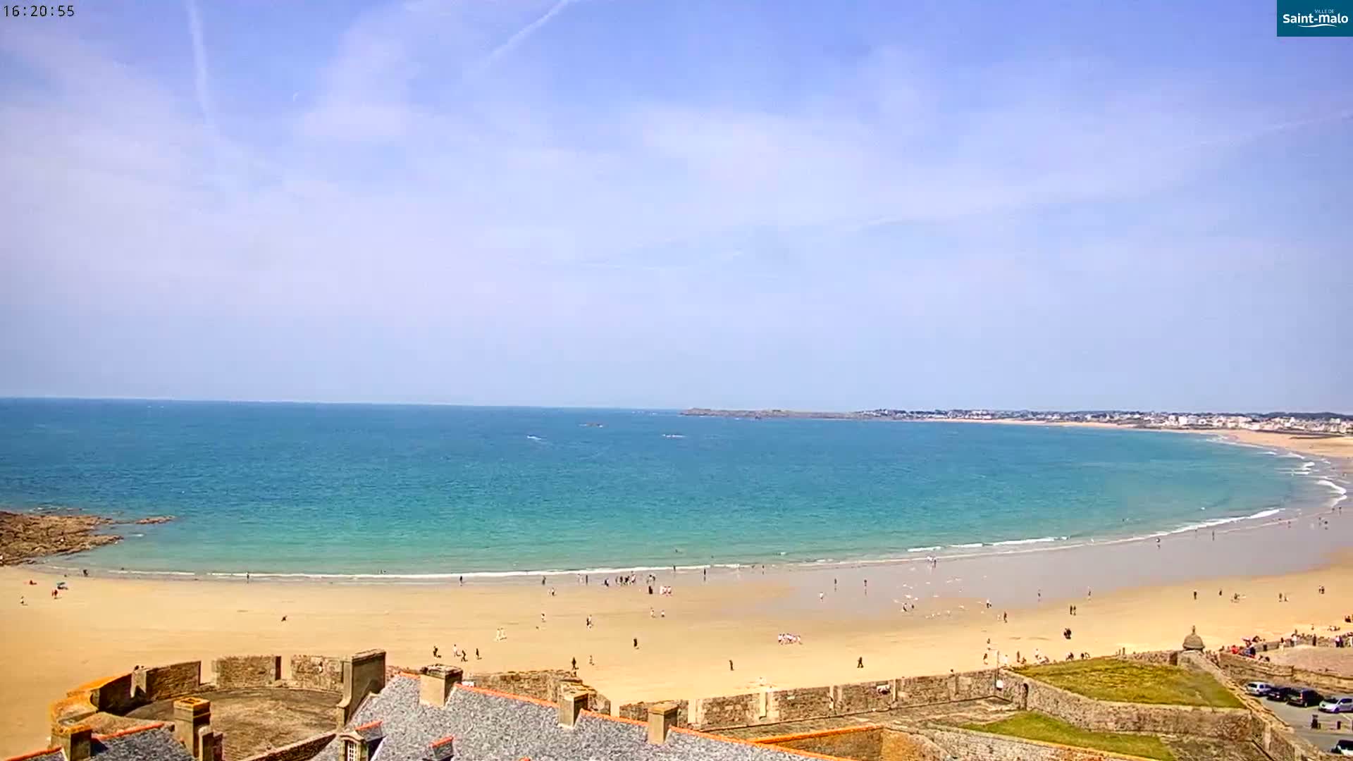 A wide sandy beach curves along a turquoise ocean under a mostly sunny sky, with a stone wall and buildings visible in the foreground.