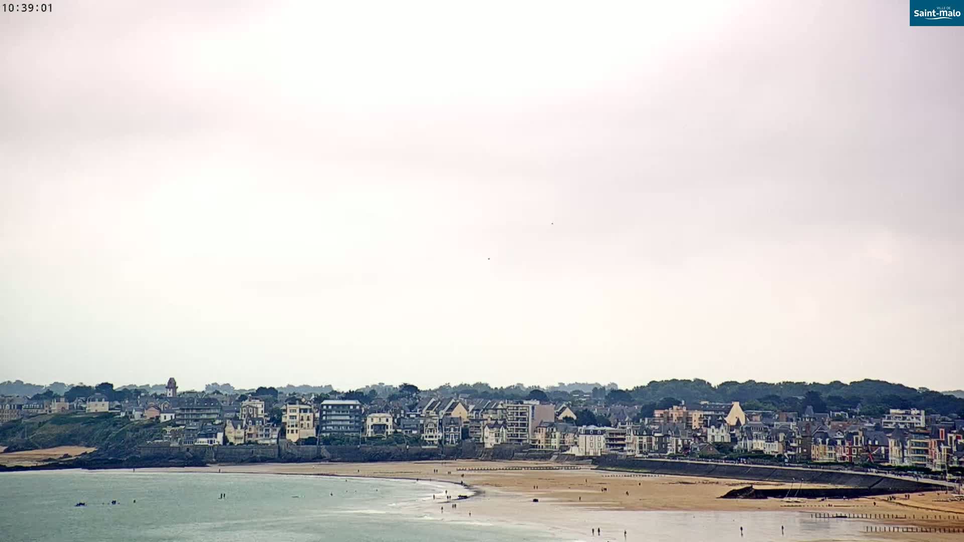 A long sandy beach stretches out in front of a town of low-rise buildings under an overcast sky.