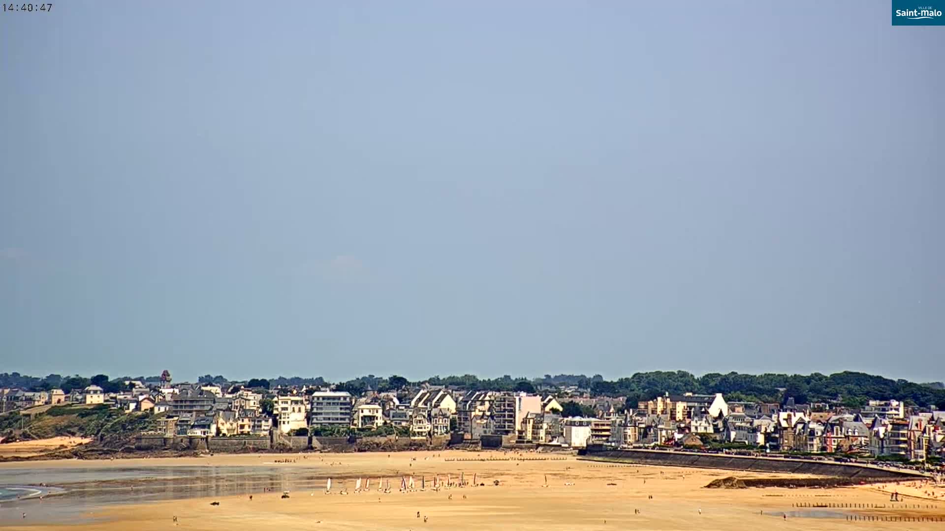 A long sandy beach stretches out before a town of buildings along the coast, under a mostly clear sky.
