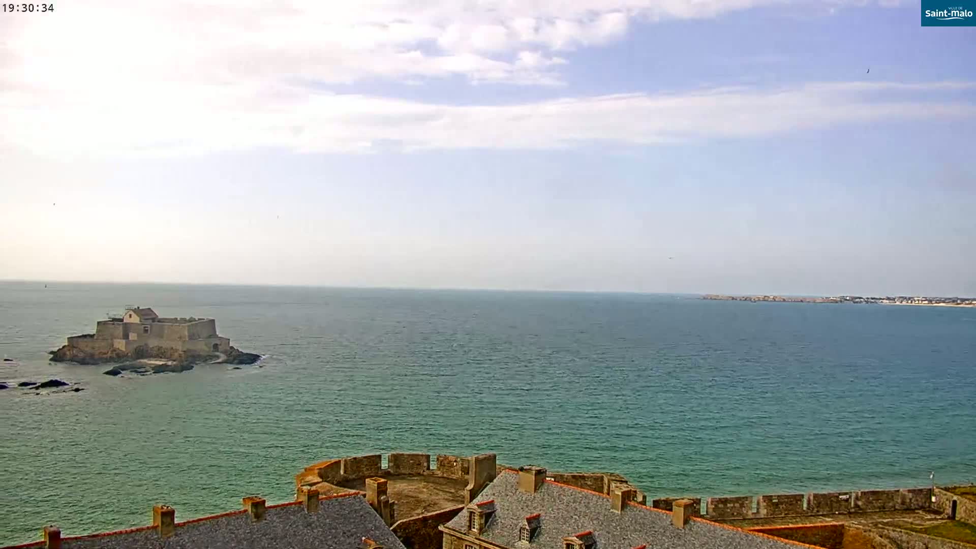 A coastal view shows a stone fortress on a small island in a calm sea under a partly cloudy sky, with another section of fortified walls and buildings in the foreground.