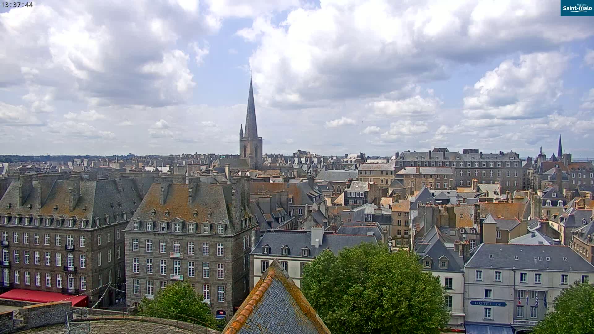 A partly cloudy day reveals a panoramic view of a European city, featuring numerous stone buildings with steeply pitched roofs, and a tall church spire in the distance.