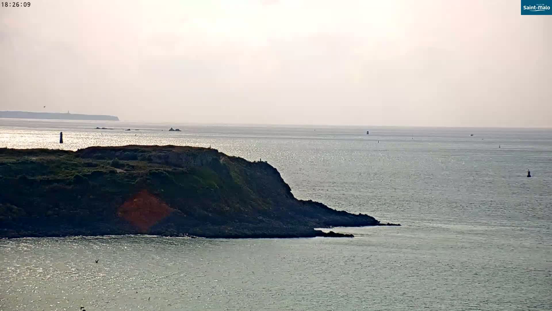 A rocky, green-covered headland juts into a calm, light-blue sea under a hazy sky.
