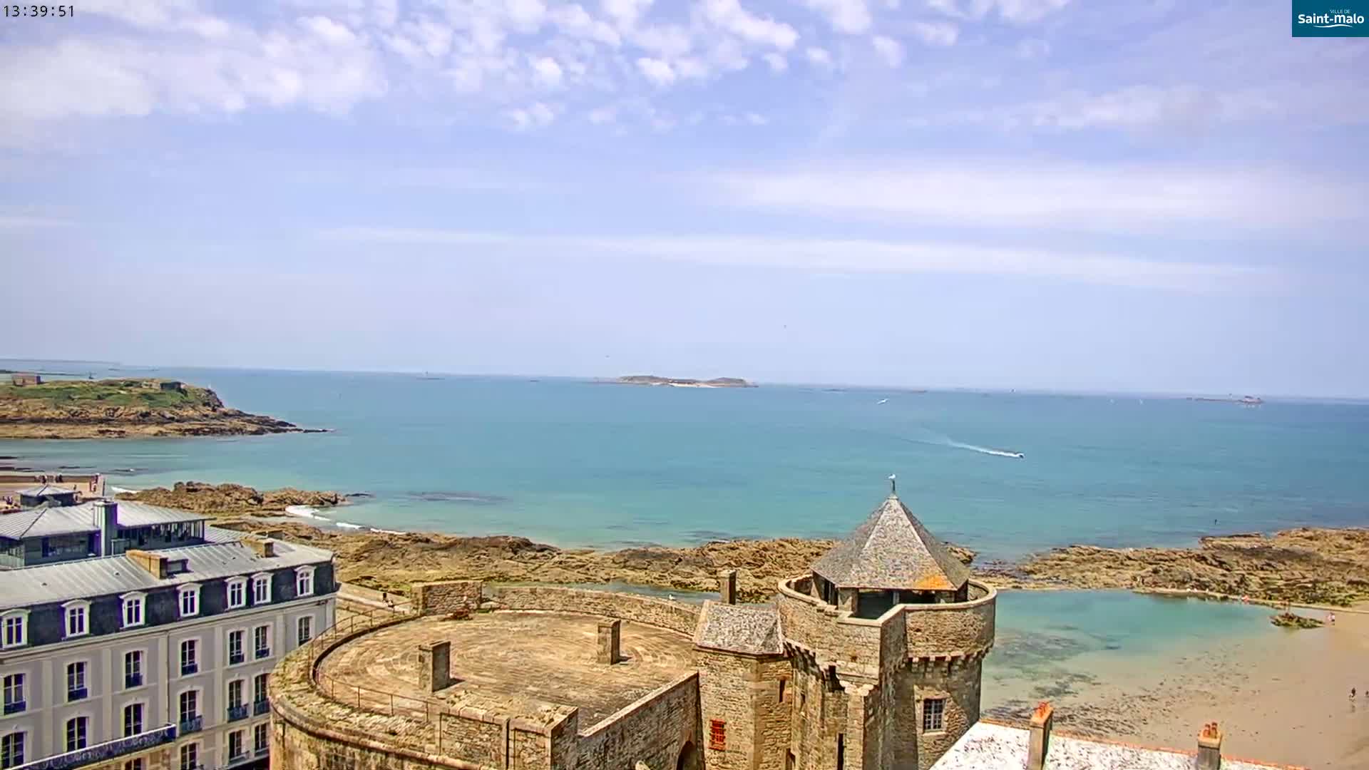 A stone castle overlooks a calm, teal ocean under a partly cloudy sky.