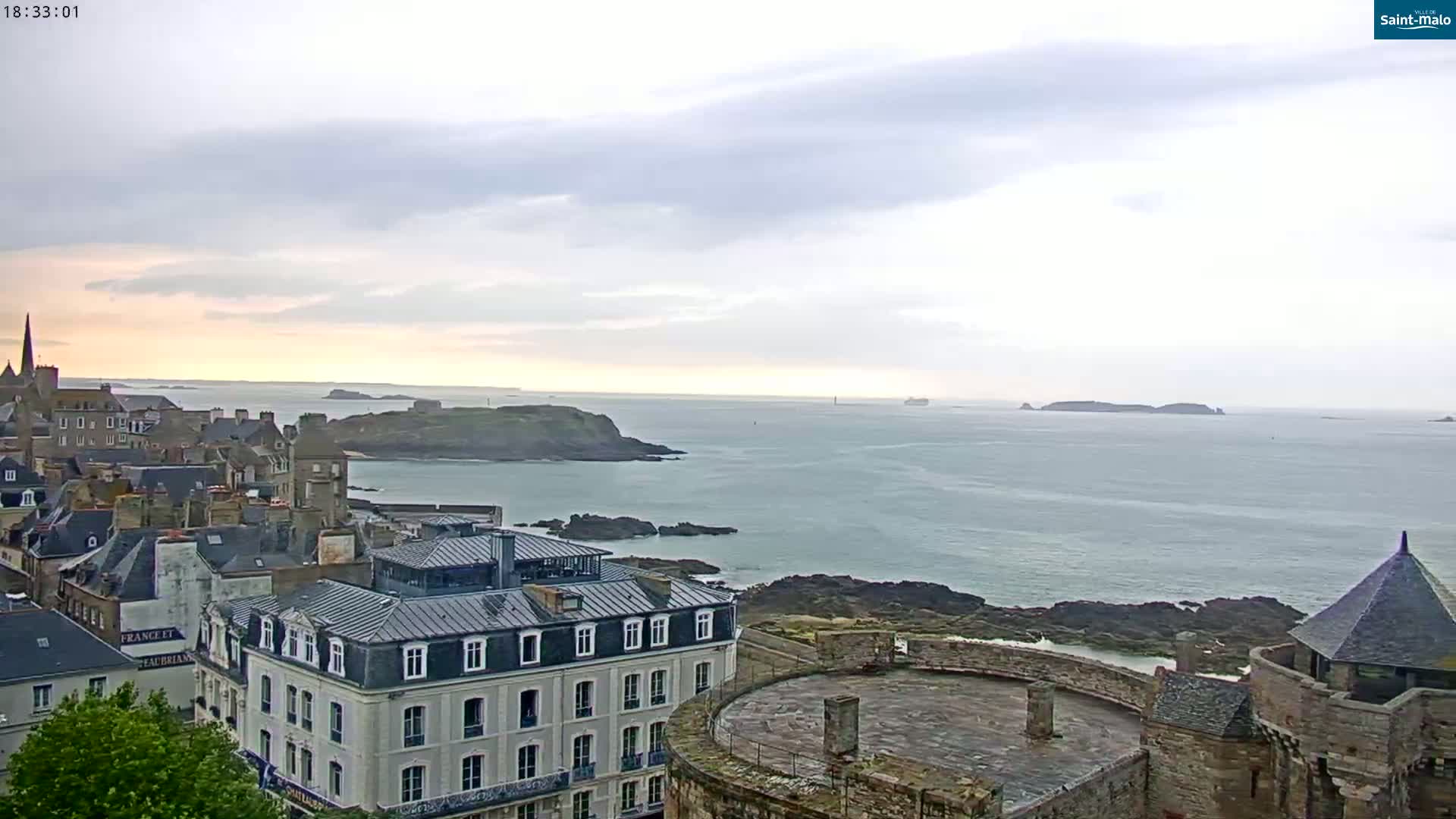A coastal town with stone buildings overlooks a calm sea under a cloudy sky.