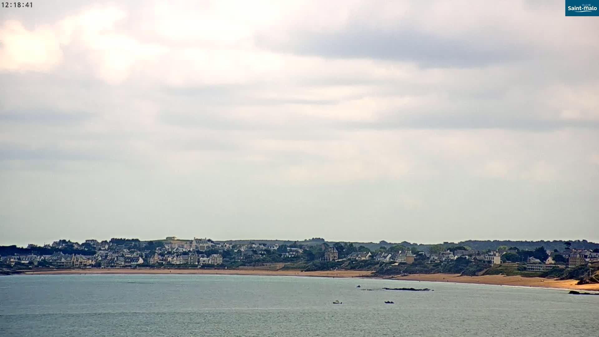 A coastal town with light-colored buildings sits on a sandy beach under a mostly cloudy sky.