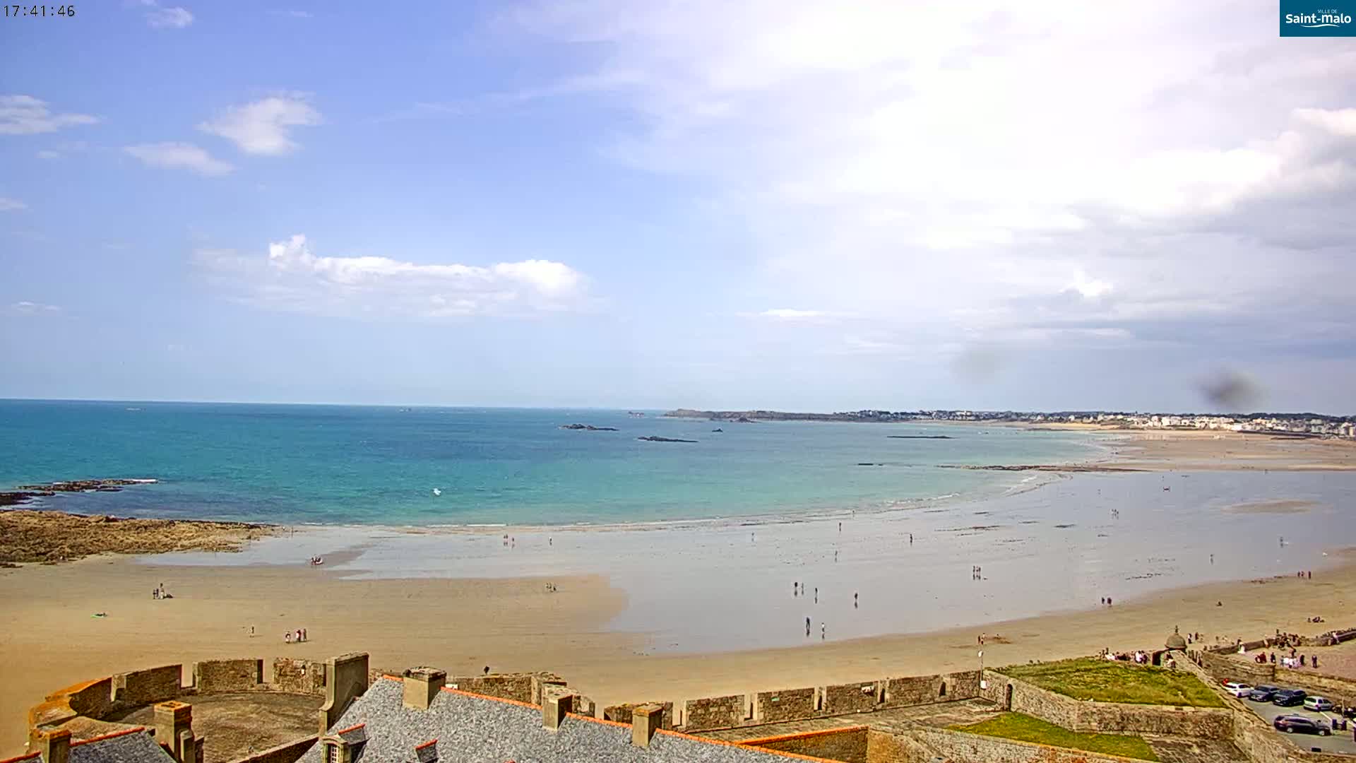 A partly cloudy sky overlooks a sandy beach and calm turquoise ocean, with a stone fortress in the foreground.