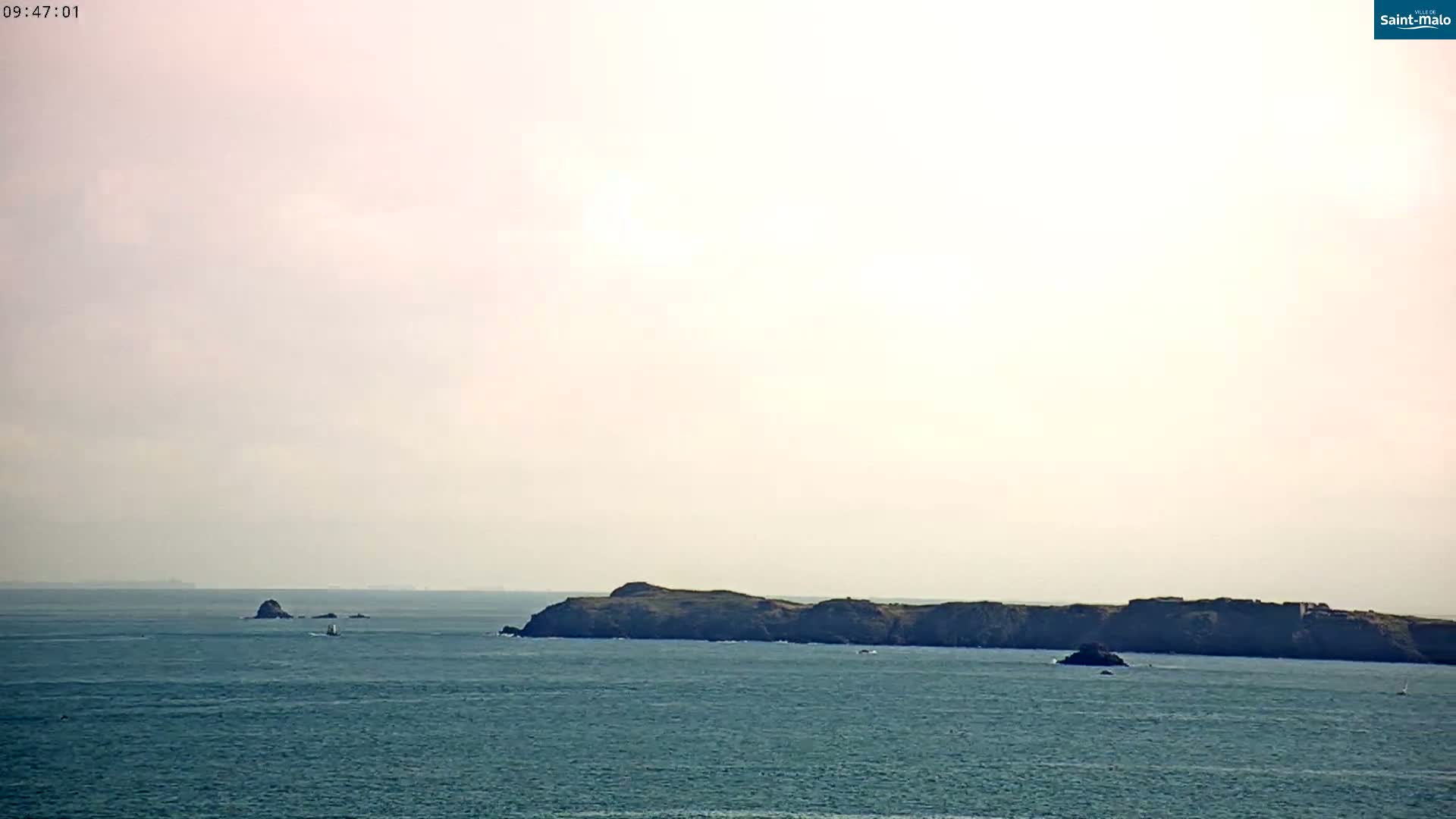 A hazy, light-colored sky overlooks a calm ocean with a dark, low-lying island in the middle distance and a smaller rock formation to the left.
