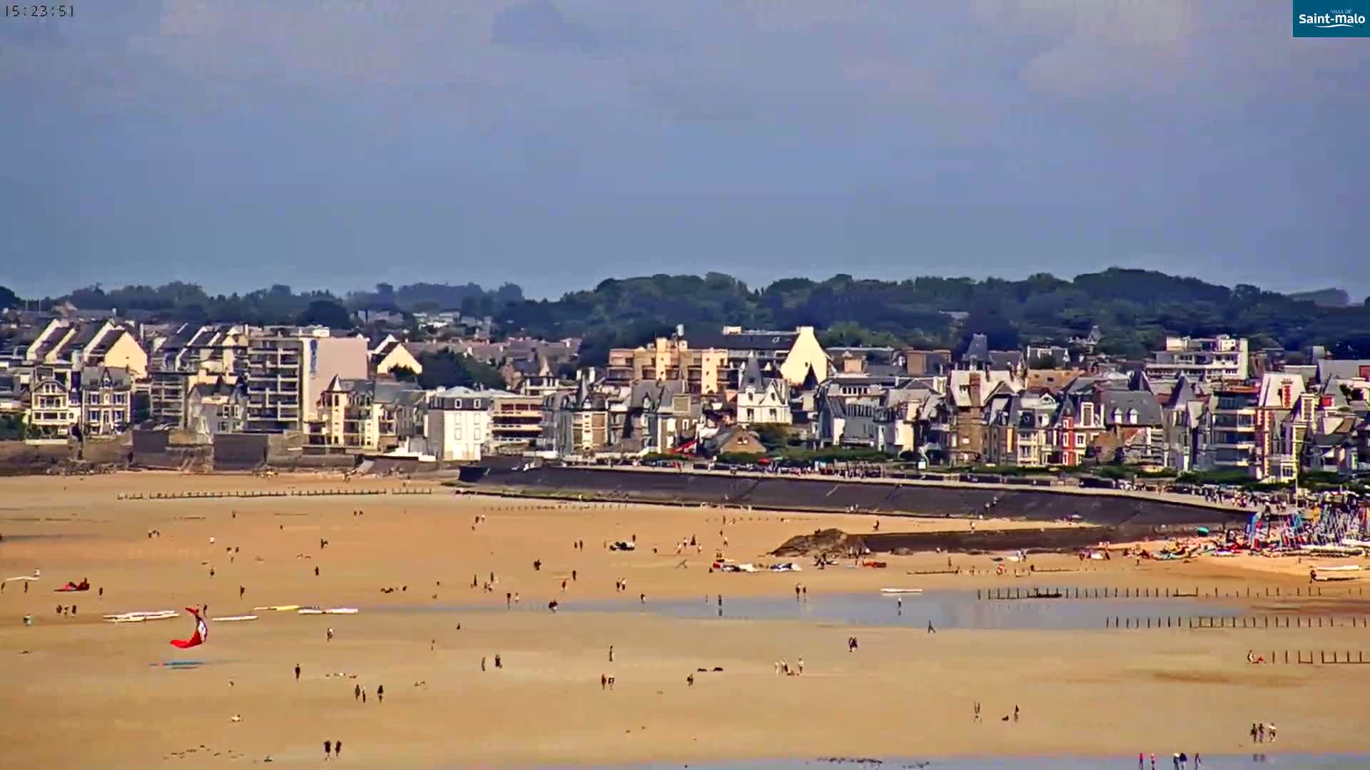 A sandy beach with scattered people and a kite surfer is in front of a town of closely-packed buildings under a mostly clear sky.
