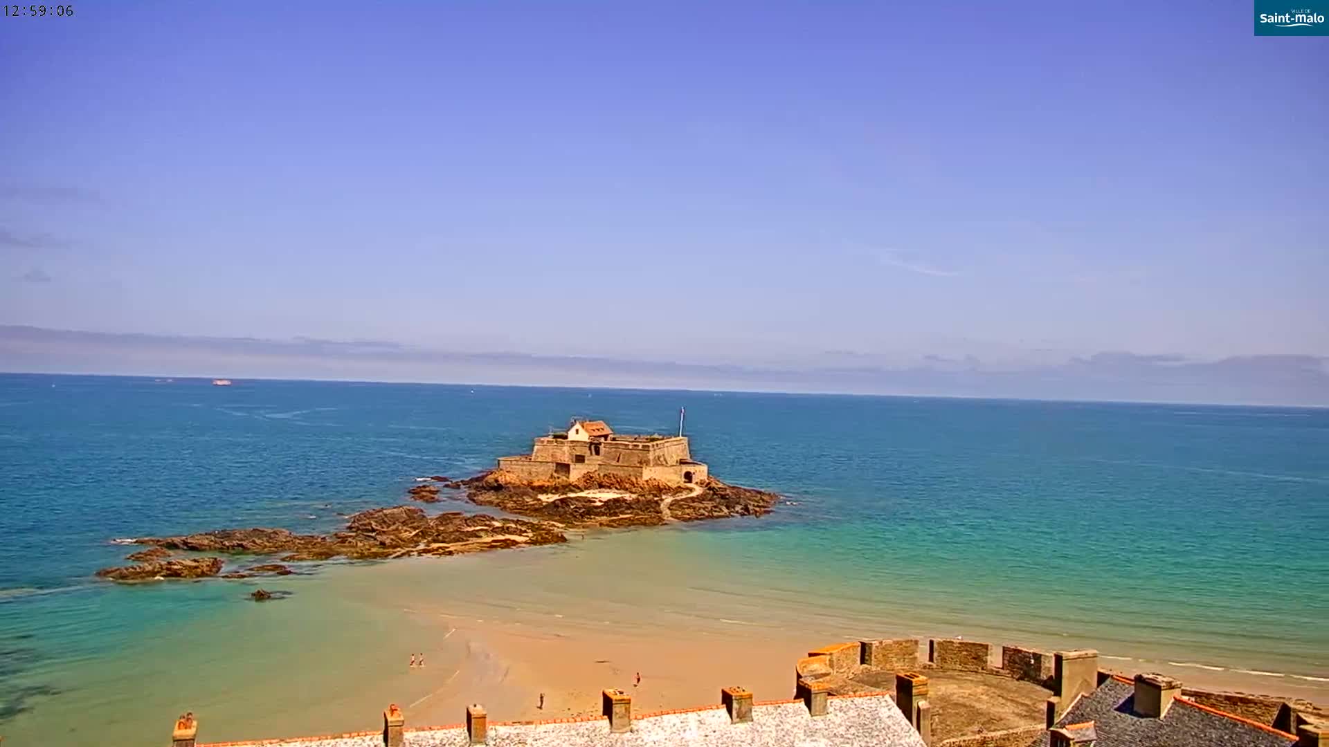 A stone fortress sits on a small island just off a sandy beach, with calm, clear blue-green water under a mostly clear, light blue sky.