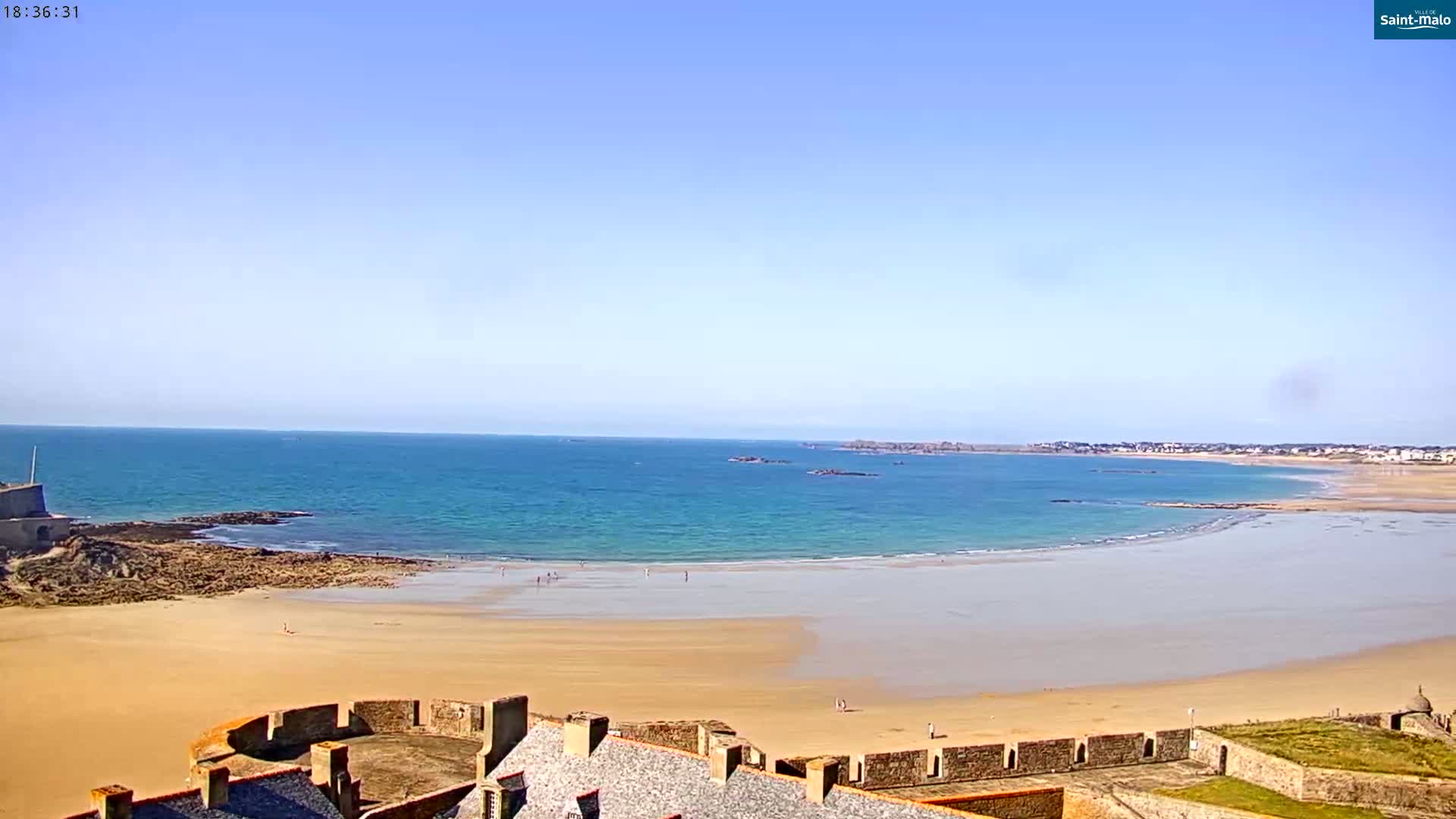A sandy beach curves around a calm, blue ocean under a clear blue sky, with a stone fortress in the foreground.