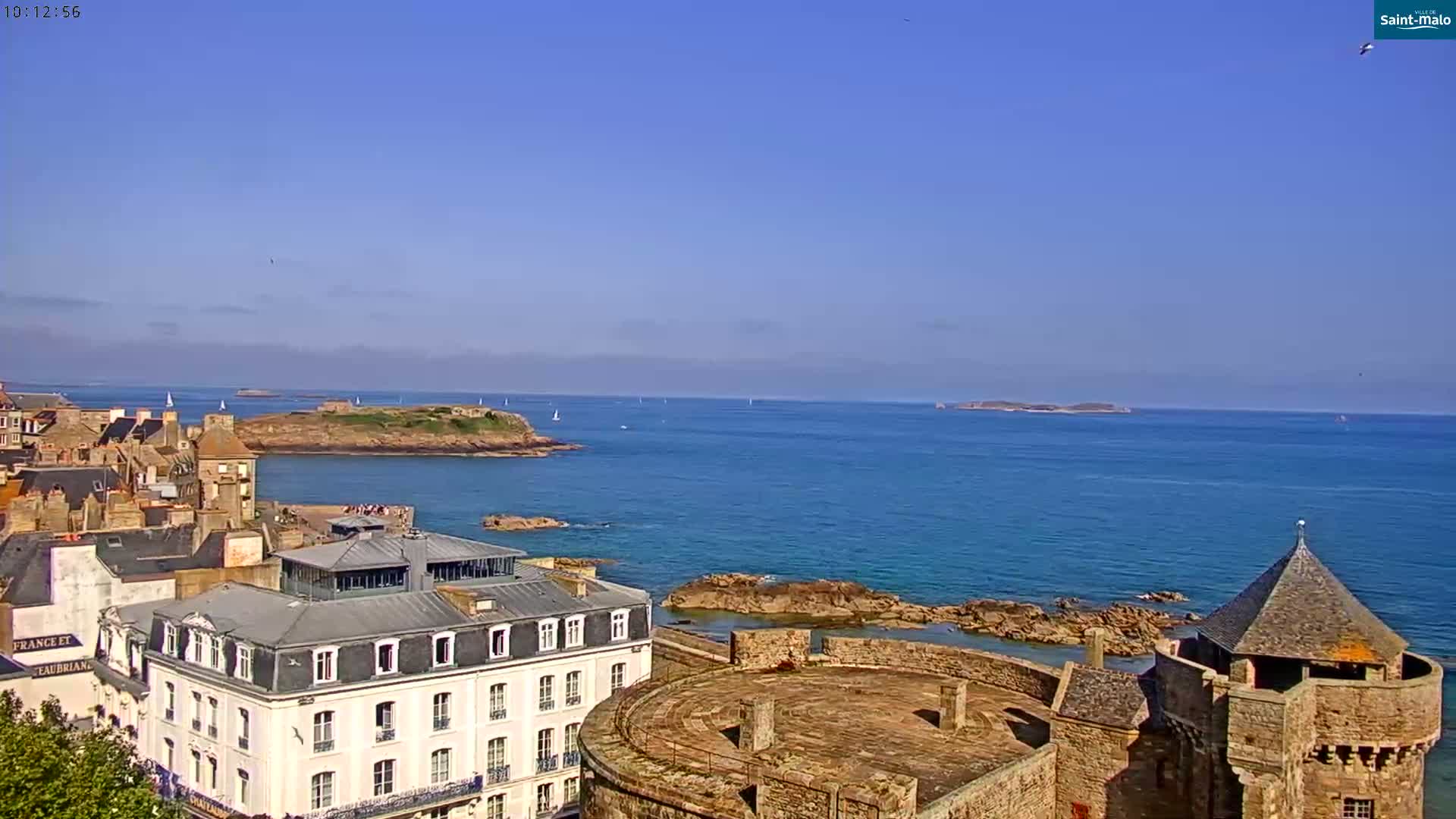 A coastal town with stone buildings, a fortress, and a clear blue sea under a bright, sunny sky.