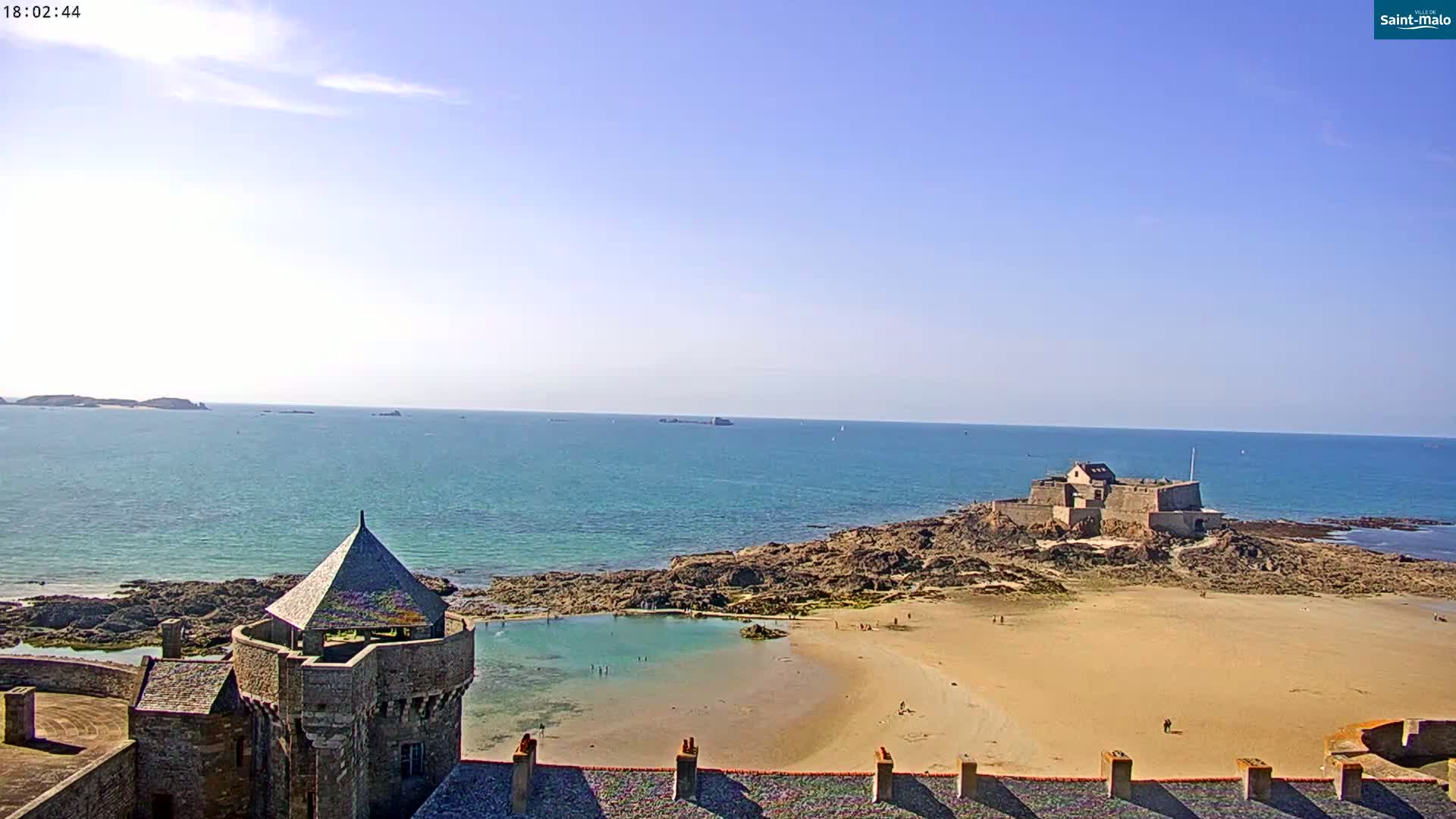 A stone coastal fortress overlooks a sandy beach and calm ocean under a clear blue sky.