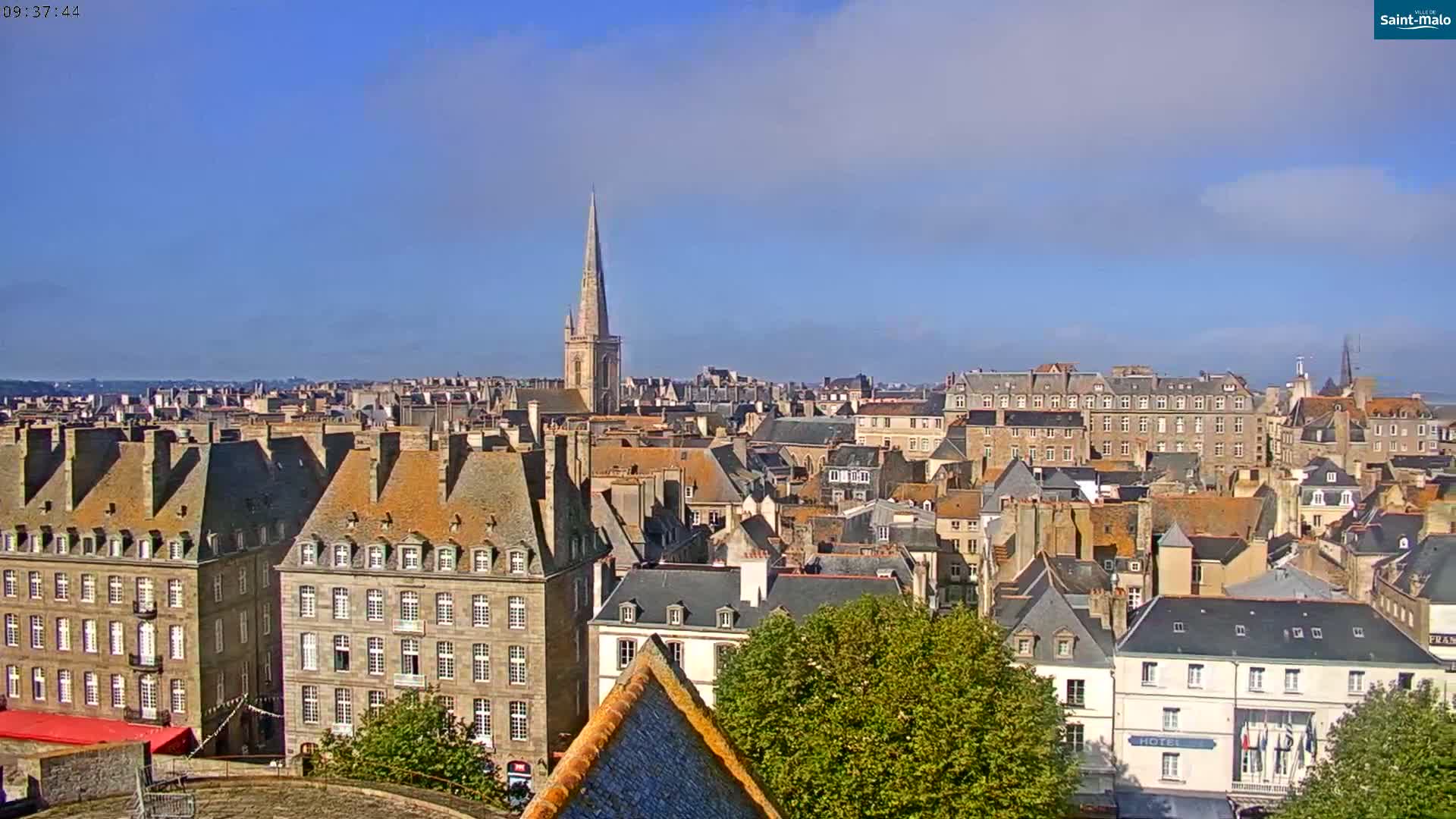 A panoramic view of a European city with predominantly gray stone buildings under a mostly clear, bright blue sky shows a church spire in the middle distance.