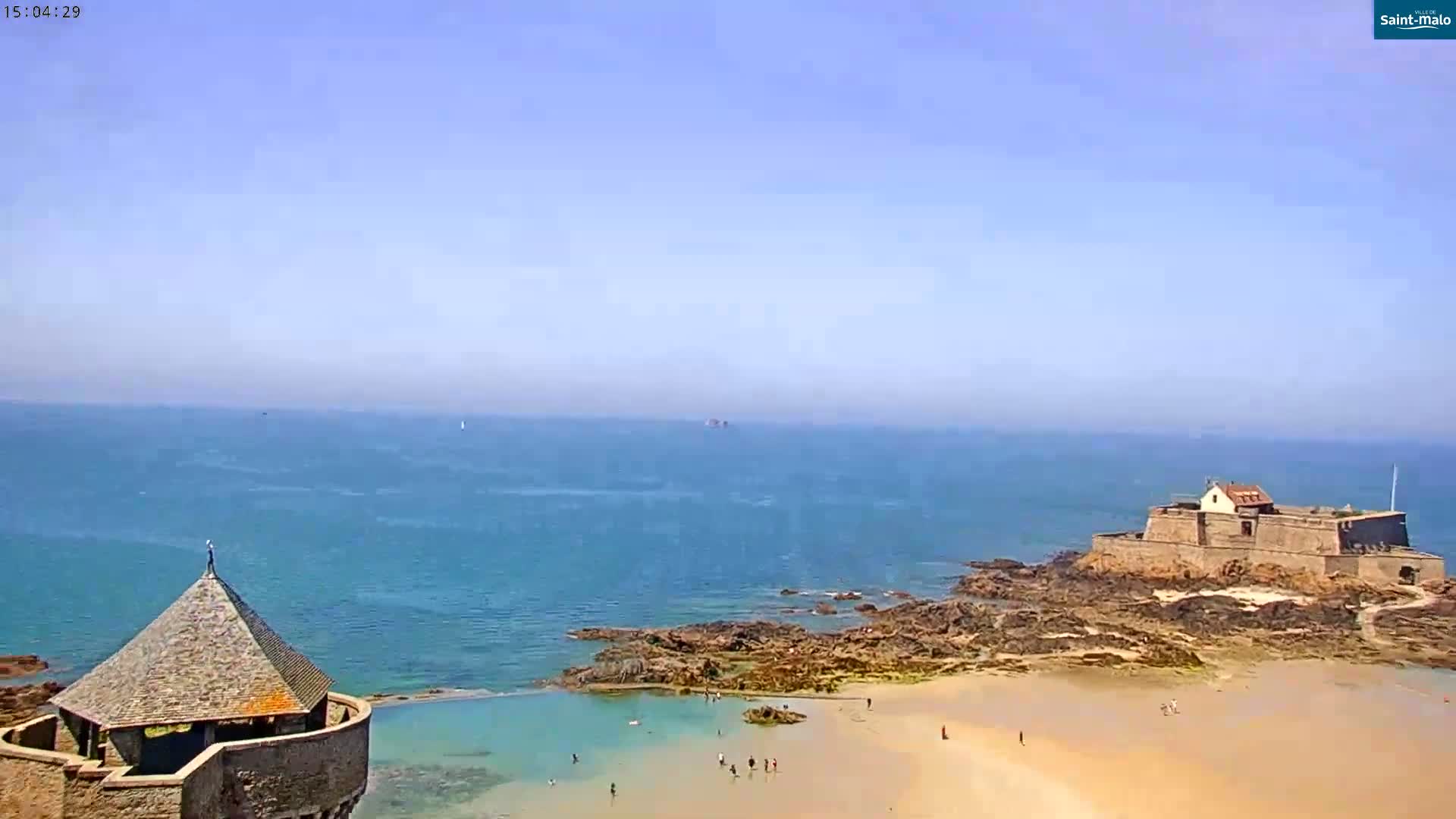 A coastal scene on a clear, sunny day shows a stone structure with a conical roof, a small sandy beach with people, rocky outcroppings, and a larger stone fortress on a nearby island in the calm ocean.