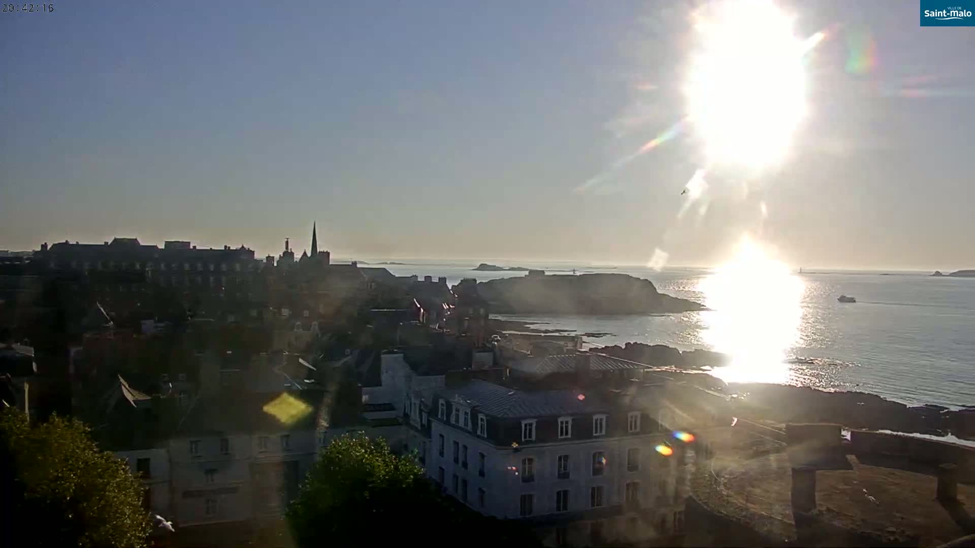 A coastal town is seen from an elevated vantage point on a sunny day, with a bright sun reflecting on the calm sea and buildings stretching out towards the water.