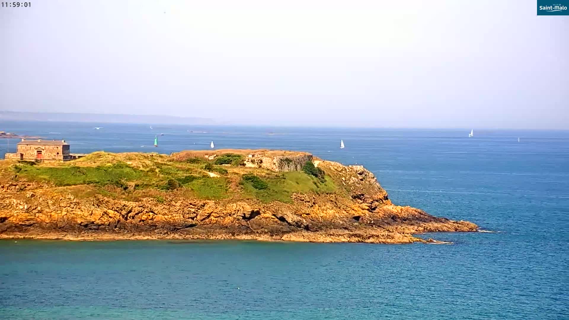 A rocky outcrop with a stone building and light green vegetation sits in a calm, blue ocean under a hazy sky, with several sailboats visible in the distance.