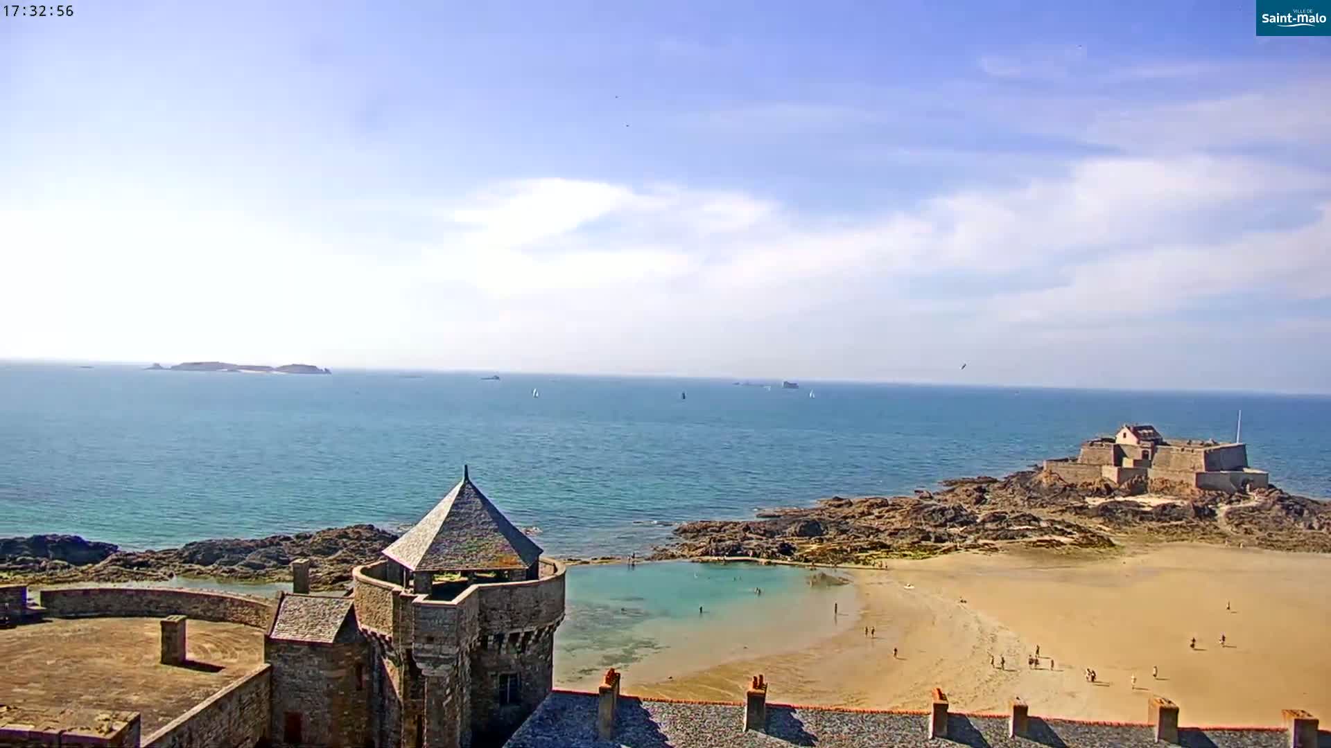 A stone castle overlooks a sandy beach and calm ocean with a distant island and a smaller fort on a rocky outcrop under a mostly sunny sky.