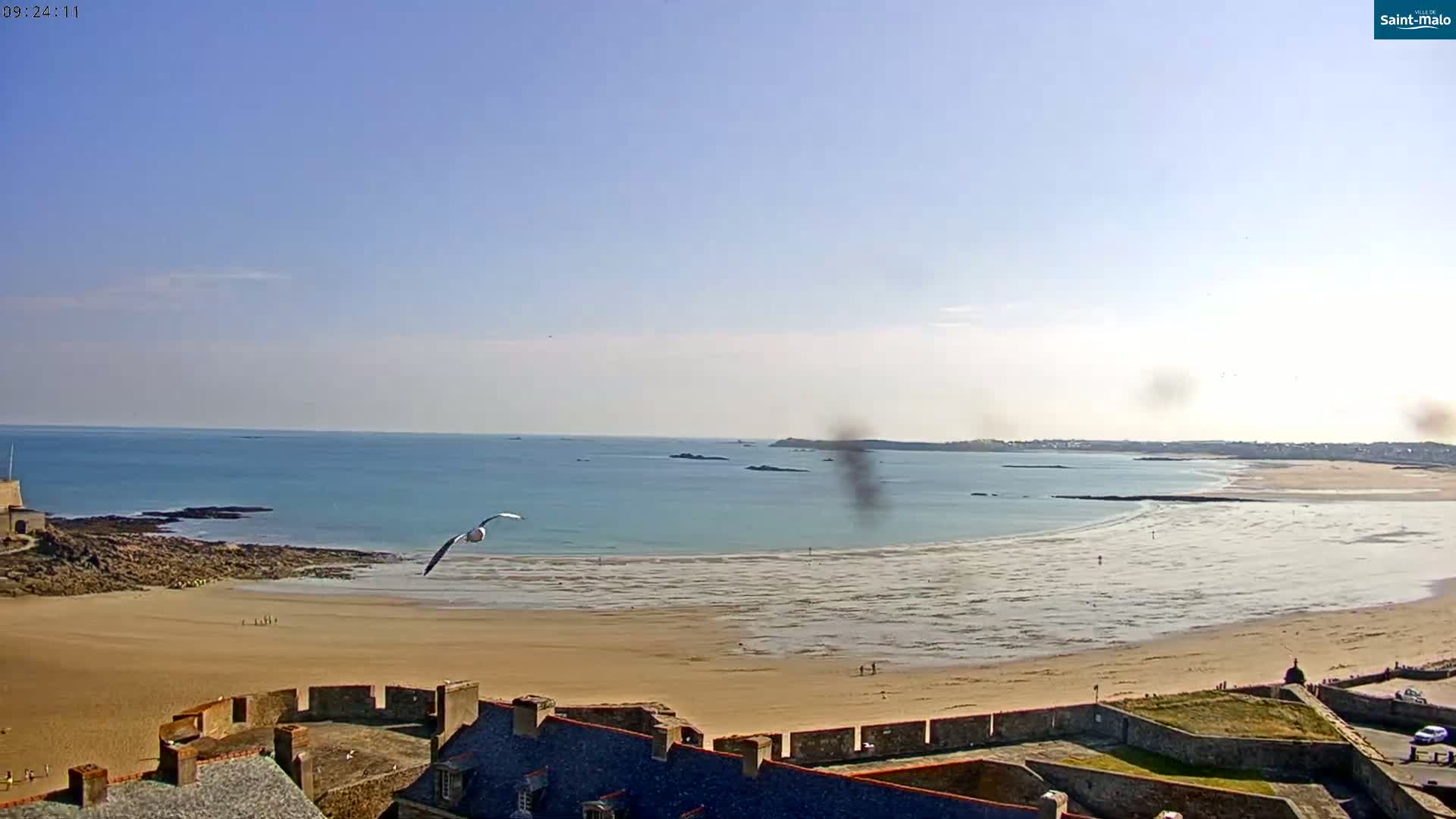 A sandy beach stretches out to a calm ocean under a clear blue sky, viewed from atop a stone fortress.