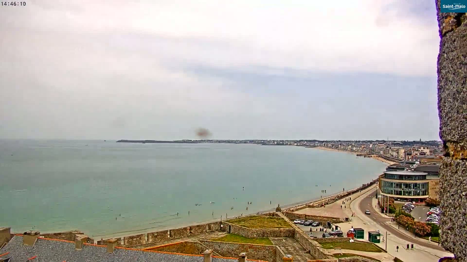 An overcast day reveals a coastal town with a sandy beach, calm sea, and stone walls overlooking the scene from a high vantage point.