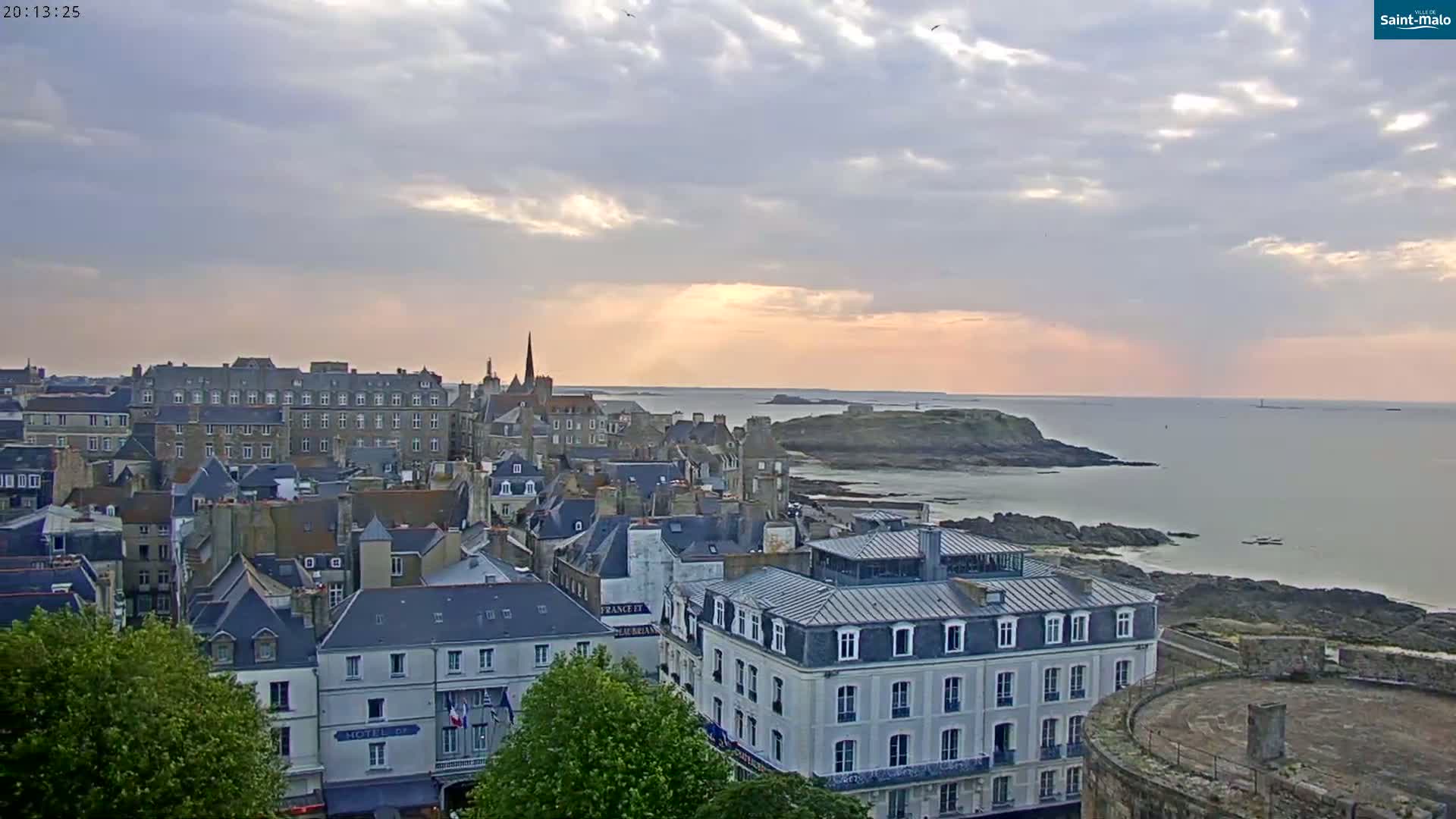 A coastal town with many gray-roofed buildings and a rocky shoreline is seen at sunset under a partly cloudy sky.