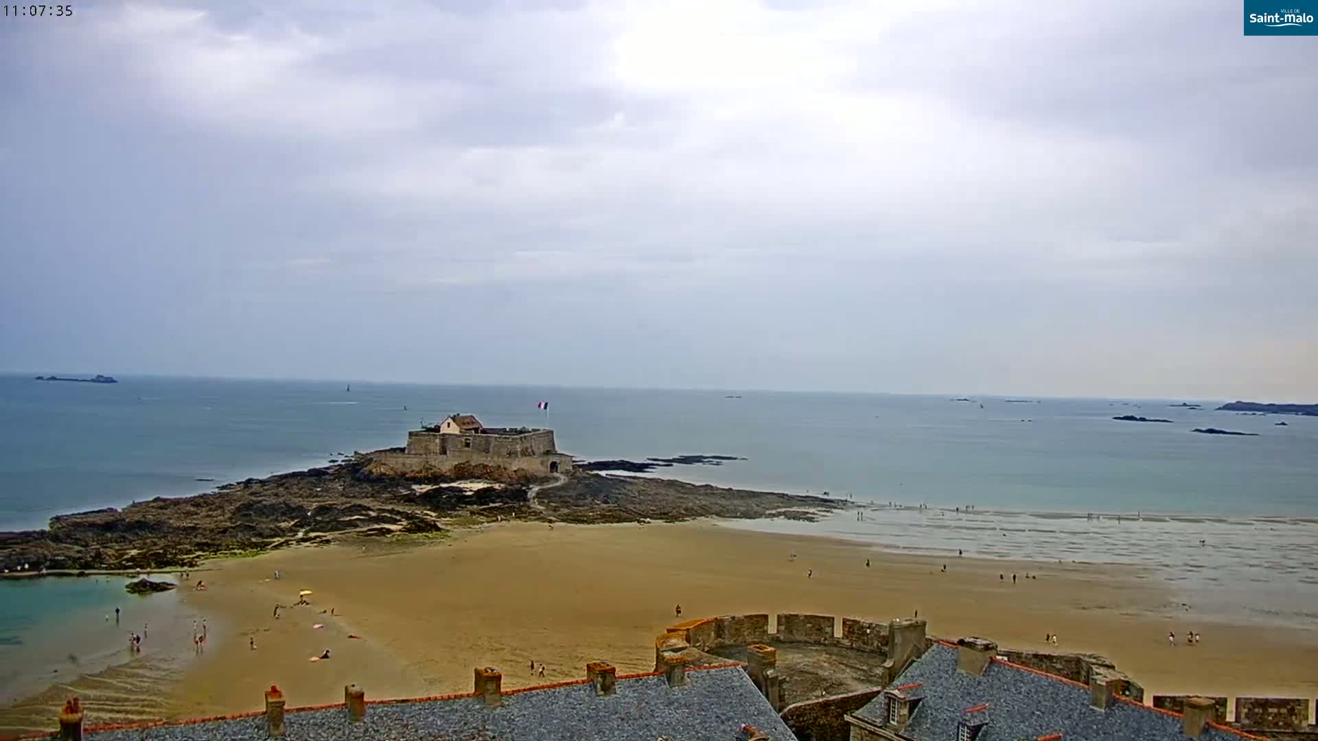A sandy beach stretches out before a stone fortress situated on a rocky outcrop, all under a cloudy sky.