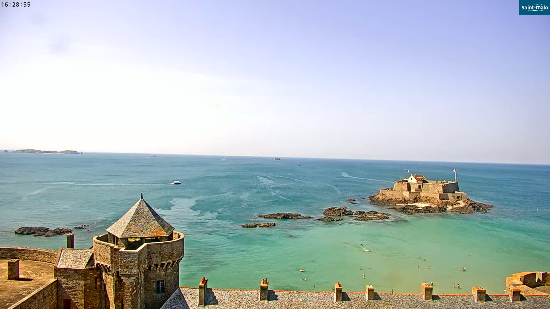 A stone castle tower overlooks a calm, teal ocean with a small island fortress and several people swimming in the shallows under a clear sky.