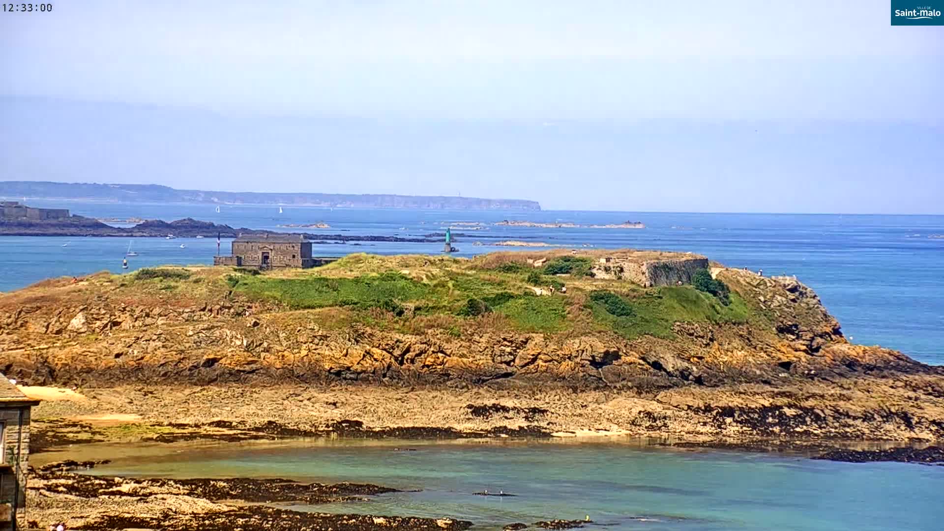 A rocky island with a stone building and green vegetation sits in a calm, blue ocean on a sunny day.