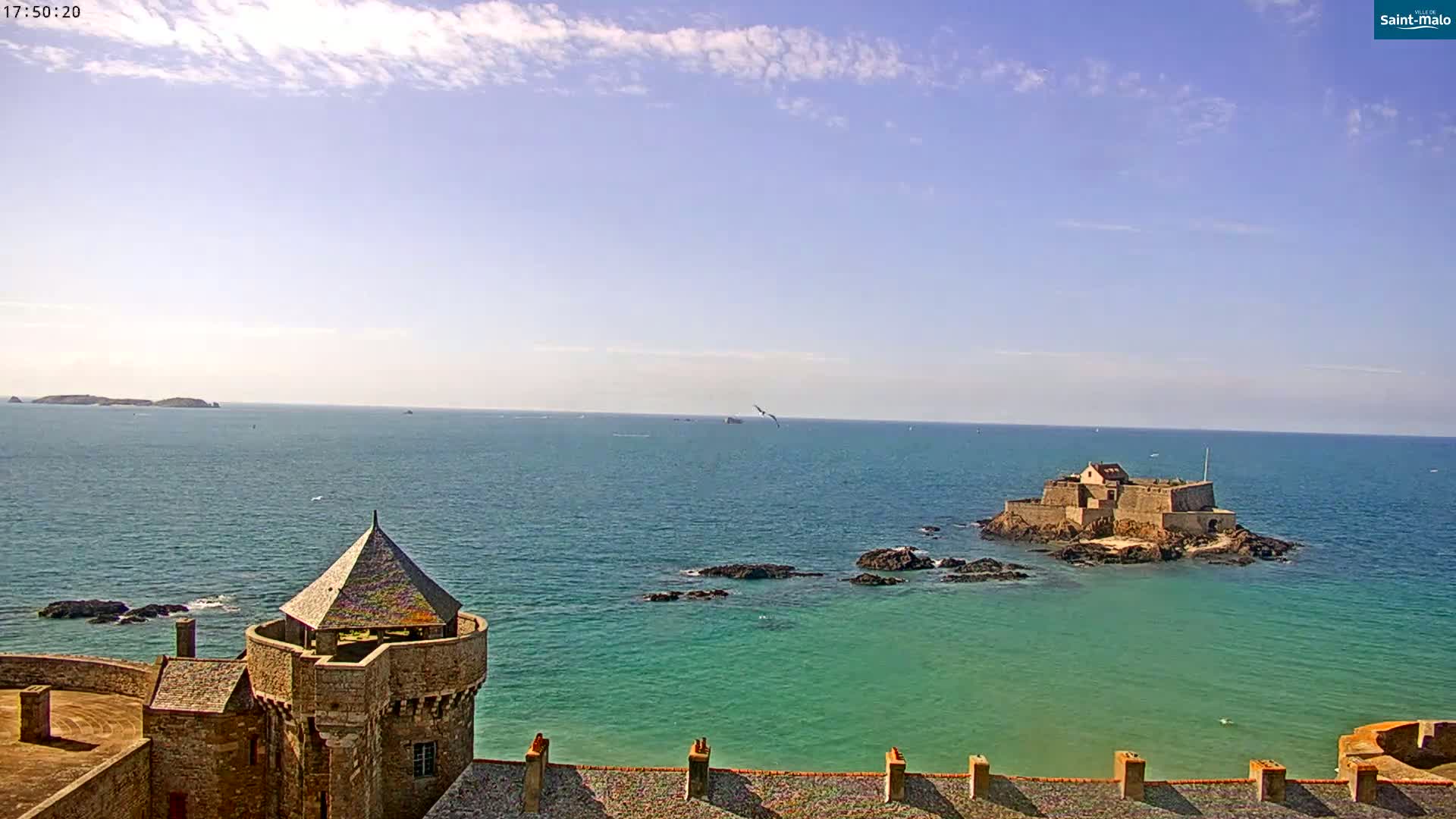 A stone tower overlooks a calm, teal ocean, with a small island fortress and distant land visible under a mostly clear sky.