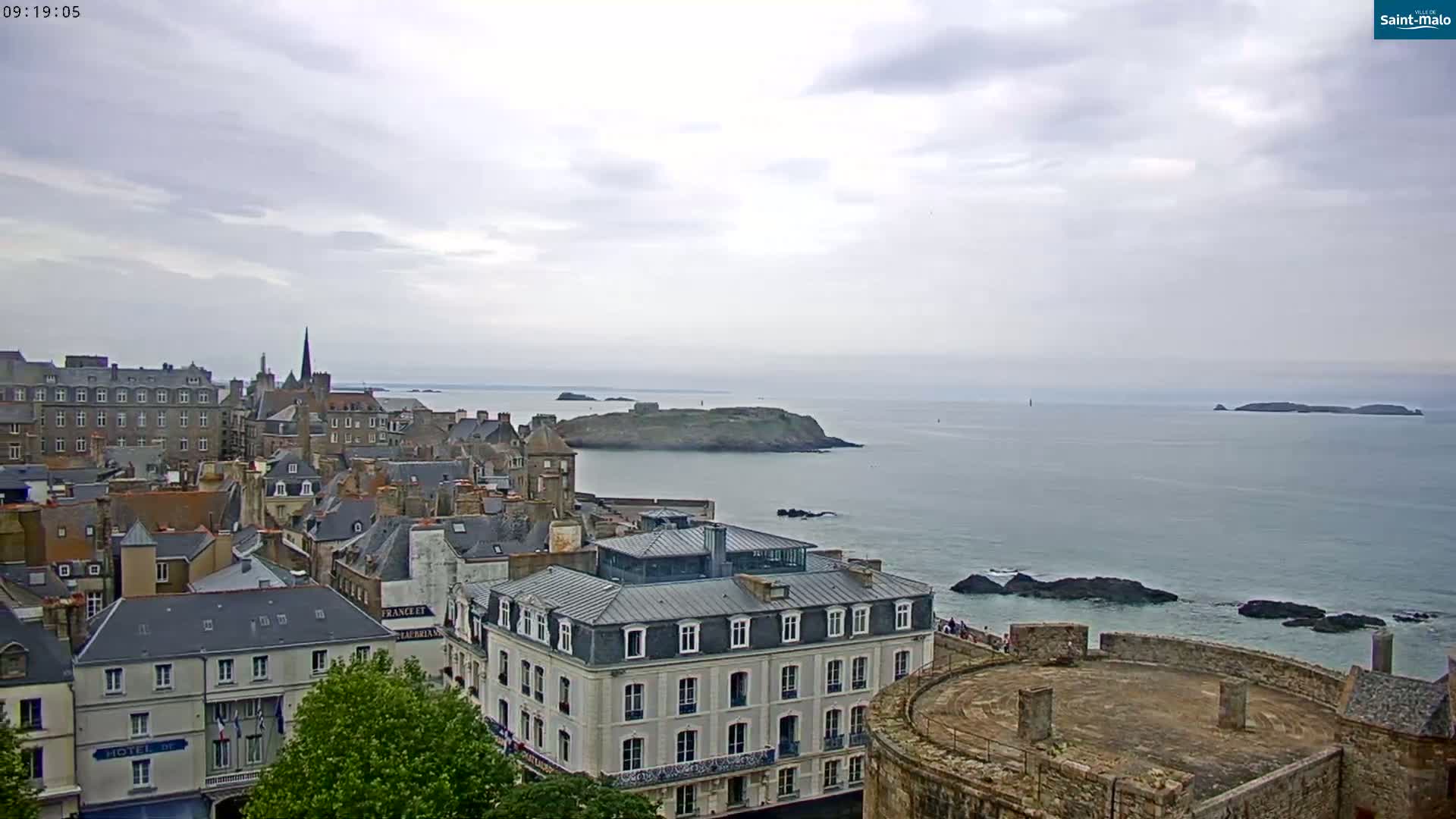 A coastal town with gray-roofed buildings overlooks a calm sea under an overcast sky.