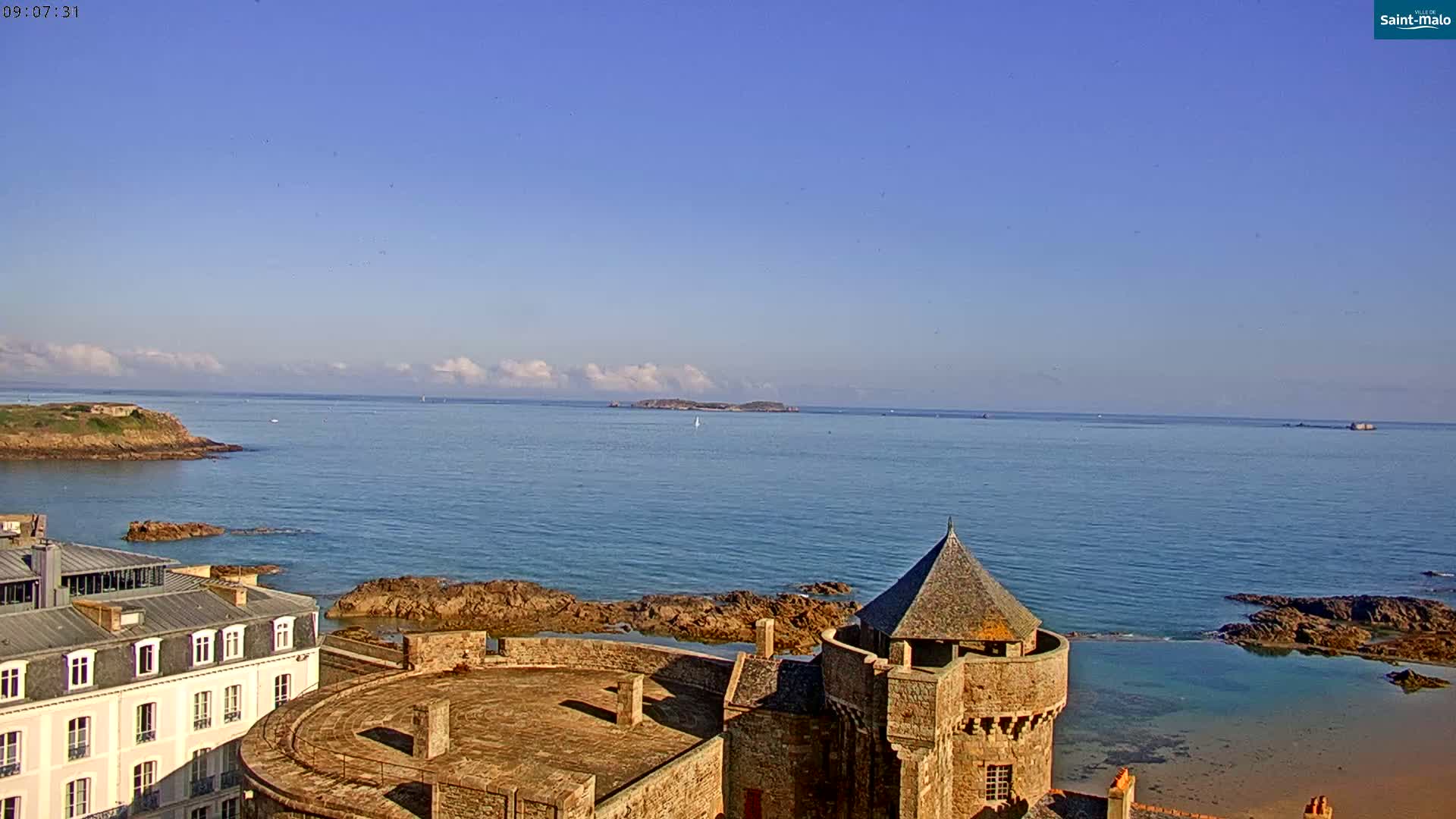 A stone castle overlooks a calm ocean under a clear blue sky with a few scattered clouds.