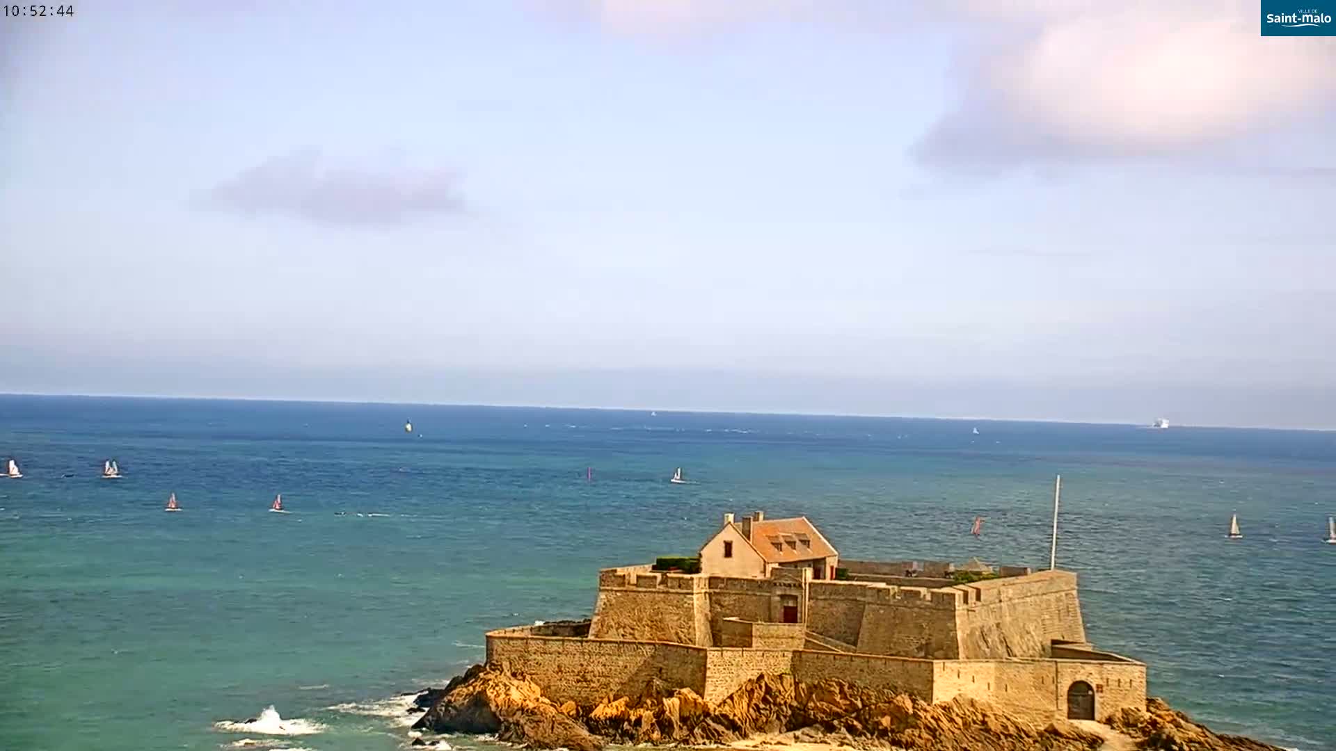 A stone fortress sits on a rocky outcropping in a calm, blue ocean under a mostly clear sky with a few scattered clouds.