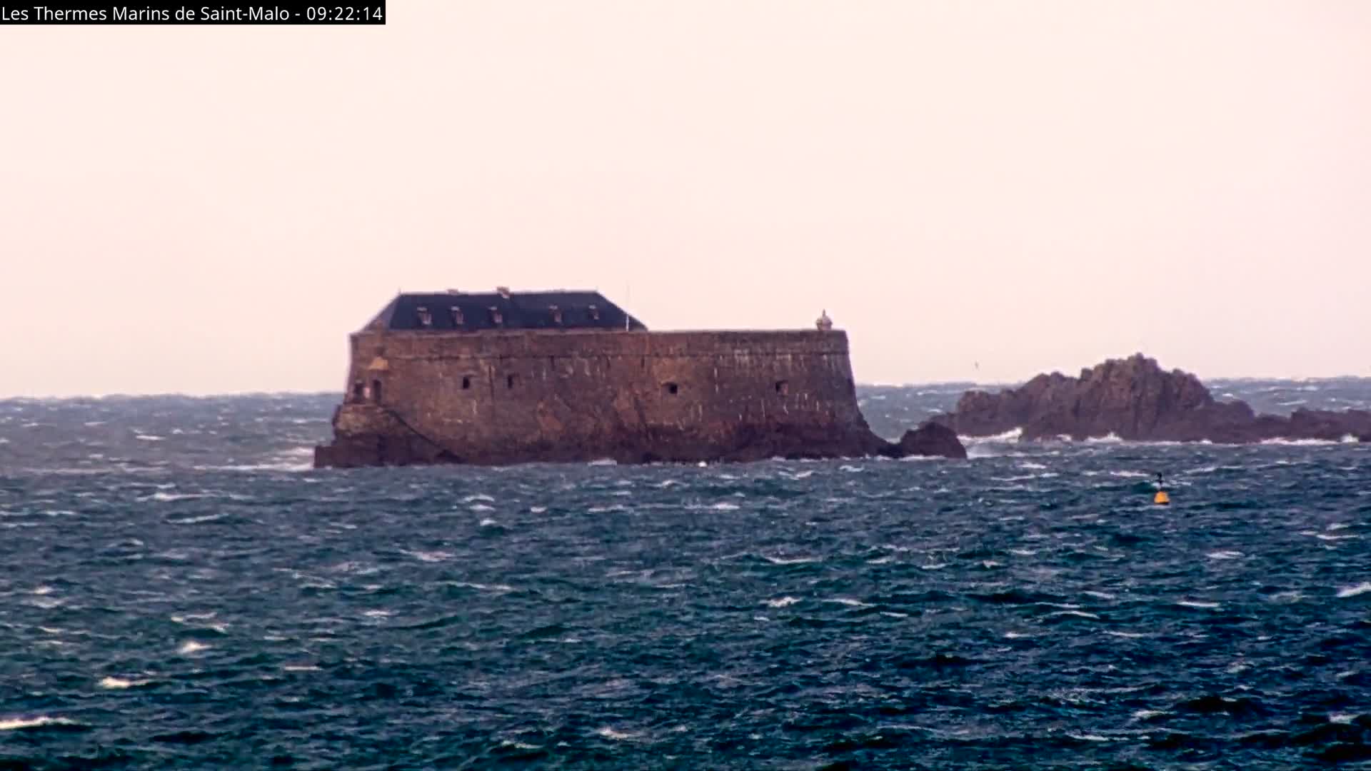 A formidable stone fort stands on a large rocky island, with smaller rocky outcrops nearby, amidst a churning, dark blue-green sea topped with whitecaps and a distant yellow buoy, under an overcast sky reflecting very windy conditions.