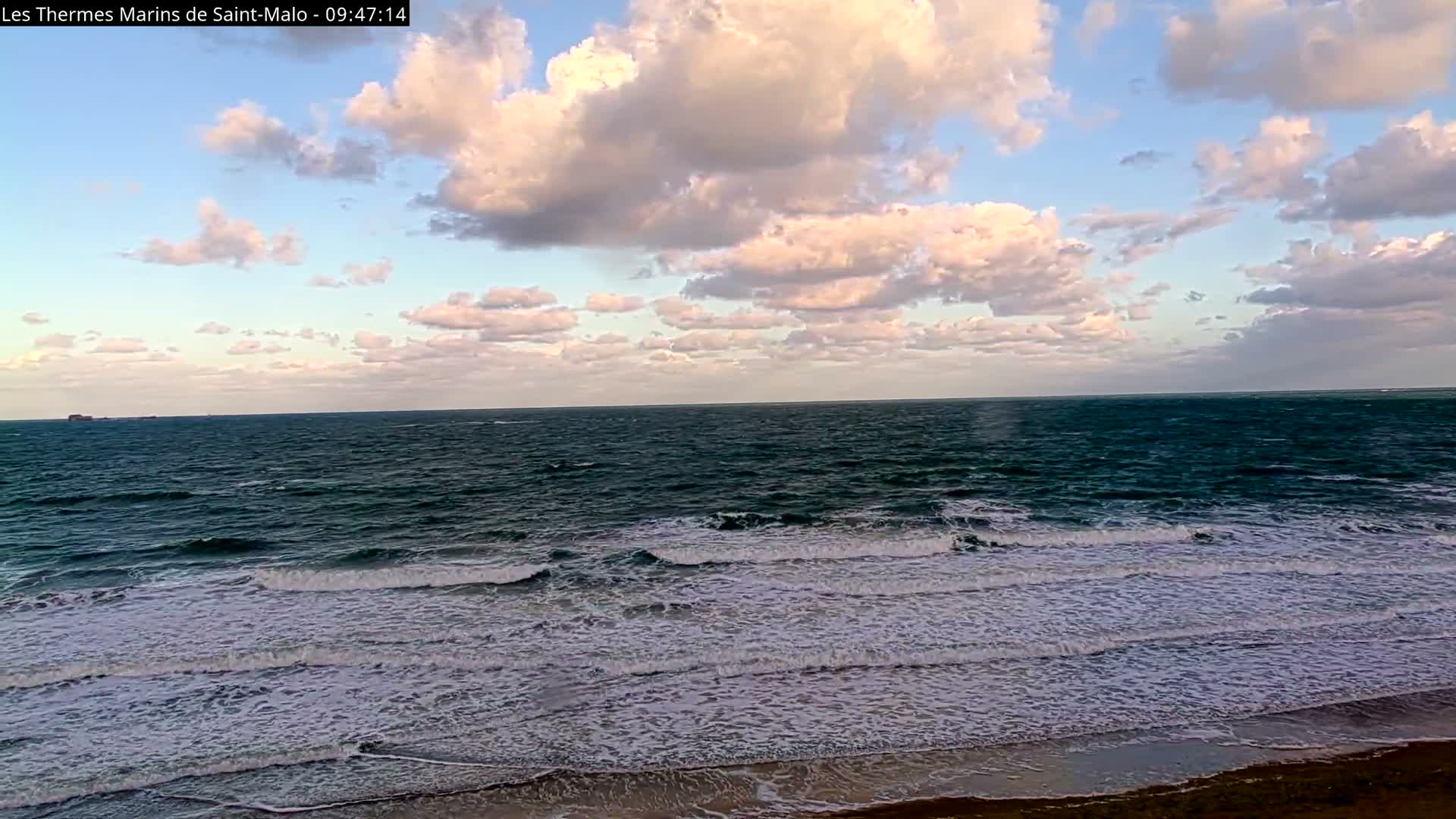 Large, frothy waves crash onto a sandy beach under a sky dotted with fluffy white clouds and patches of blue, with a distant ship visible on the horizon.